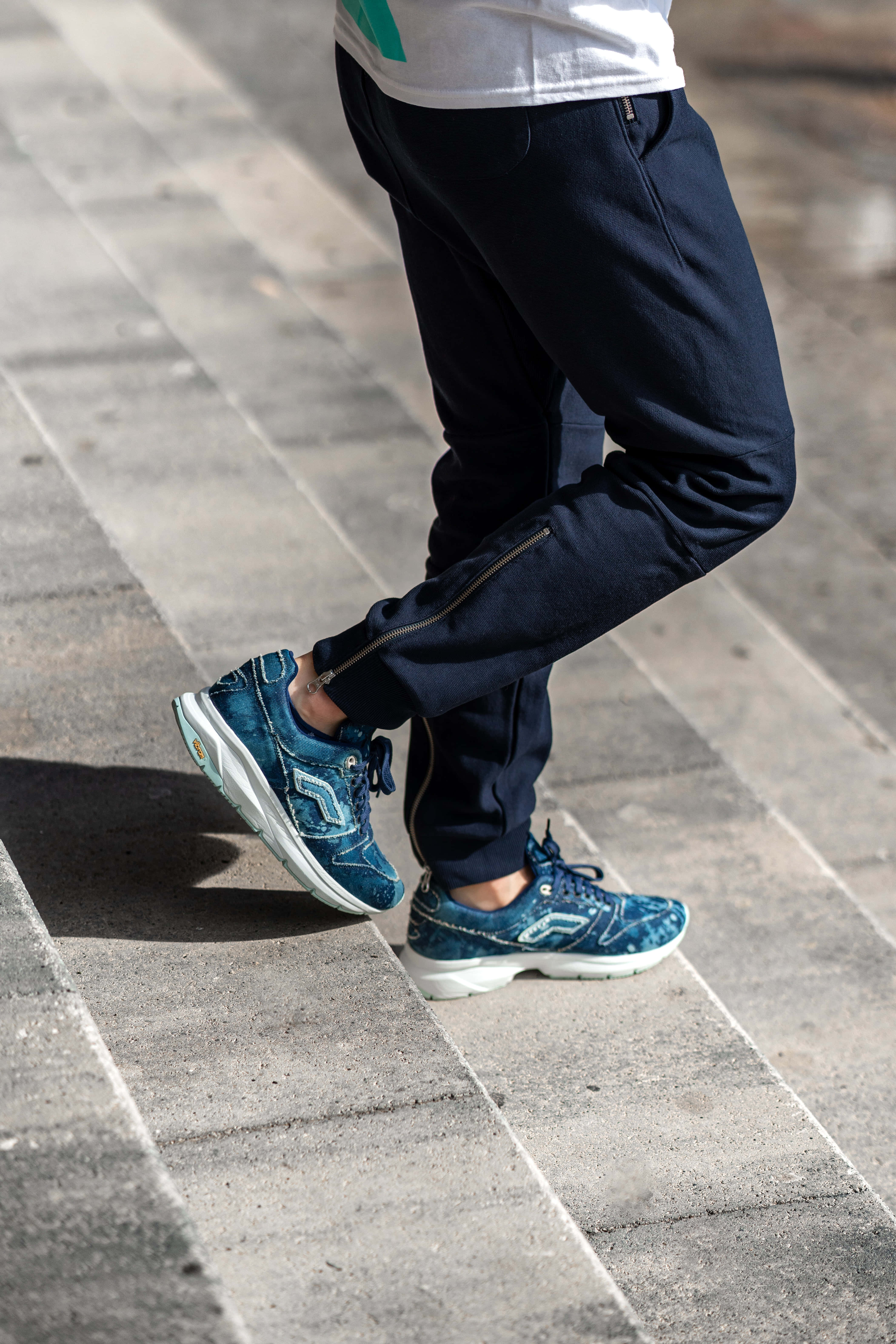 Navy Blue Shoes 桌布 壁紙 — 一個人穿著海軍藍慢跑褲和藍色運動鞋的雙腳踩在灰色石鋪路面上的特寫。