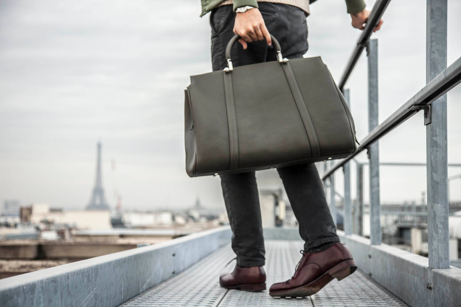 Mens Fashion 桌布 壁紙 — 一個人穿著深色褲子和酒紅色鞋子，拿著一個綠色皮革公文包，走在金屬屋頂上，俯瞰巴黎，遠處有艾菲爾鐵塔。