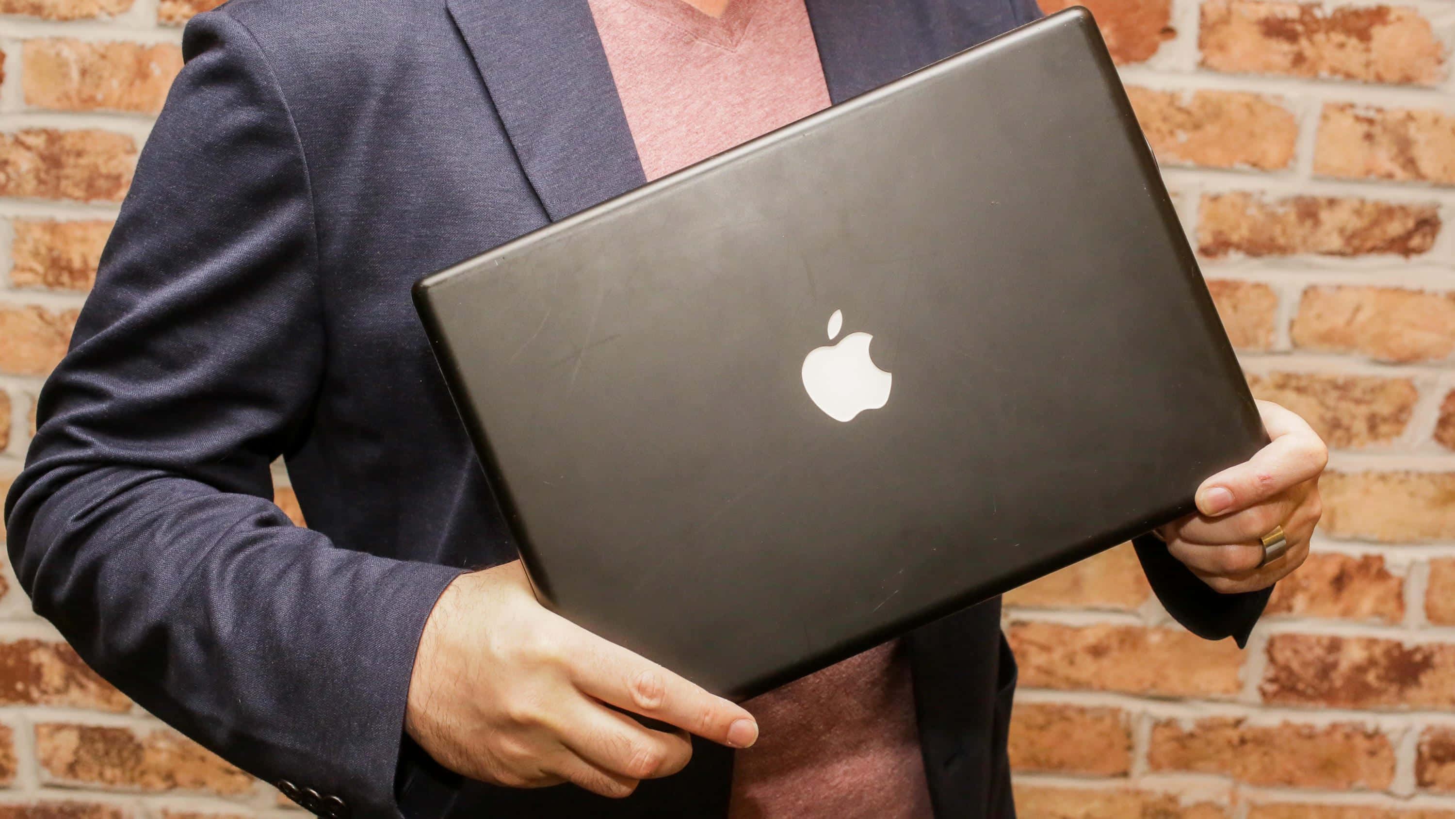 Black Macbook 桌布 壁紙 — 一個人穿著深色西裝外套，拿著黑色Apple筆記型電腦，背景是質樸的磚牆。