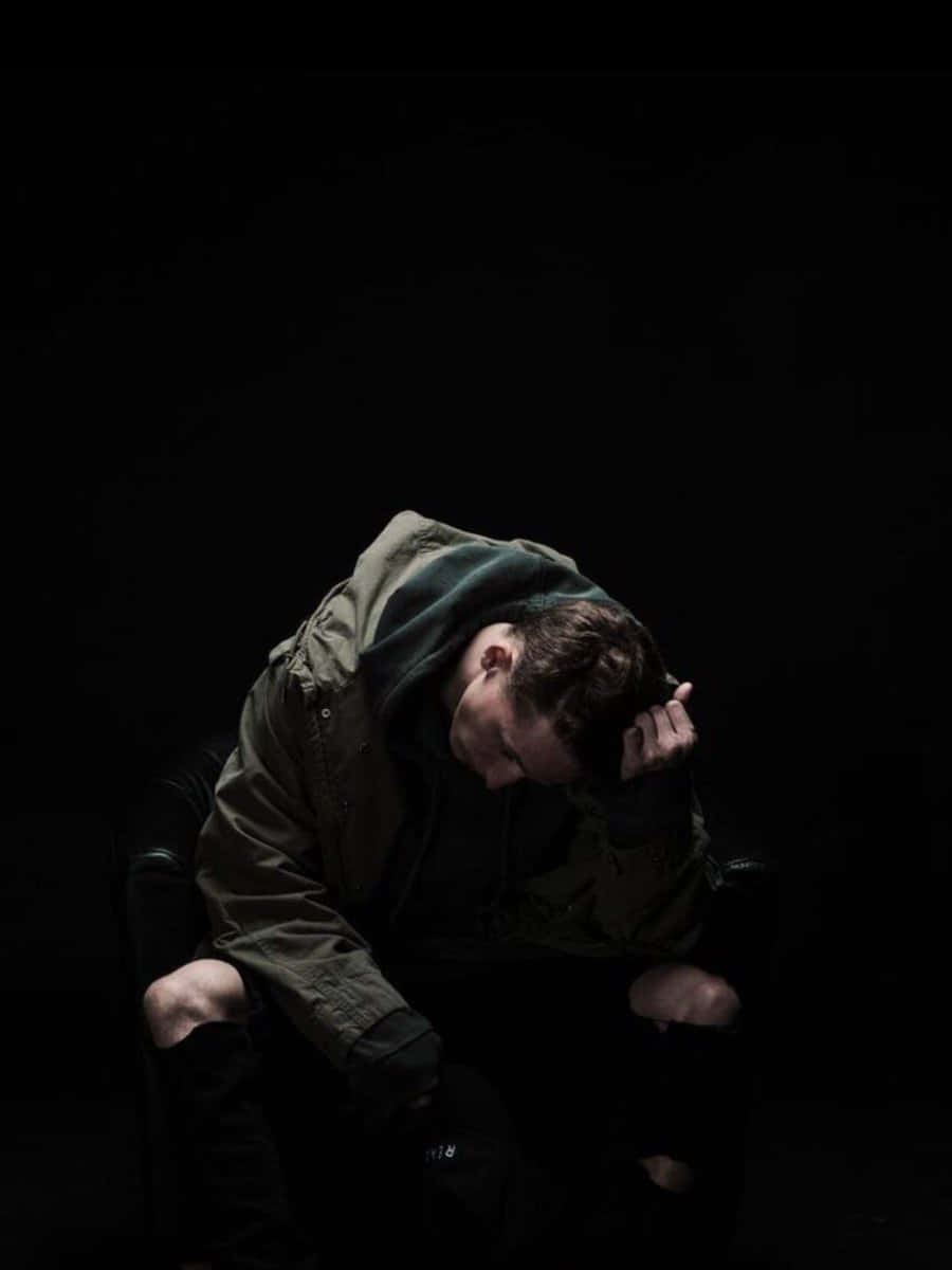 Nf Rapper 桌布 壁紙 — 一個人穿著深色連帽衫，彎腰坐在陰影中，低著頭，手放在太陽穴上，營造出陰鬱的攝影棚氛圍。