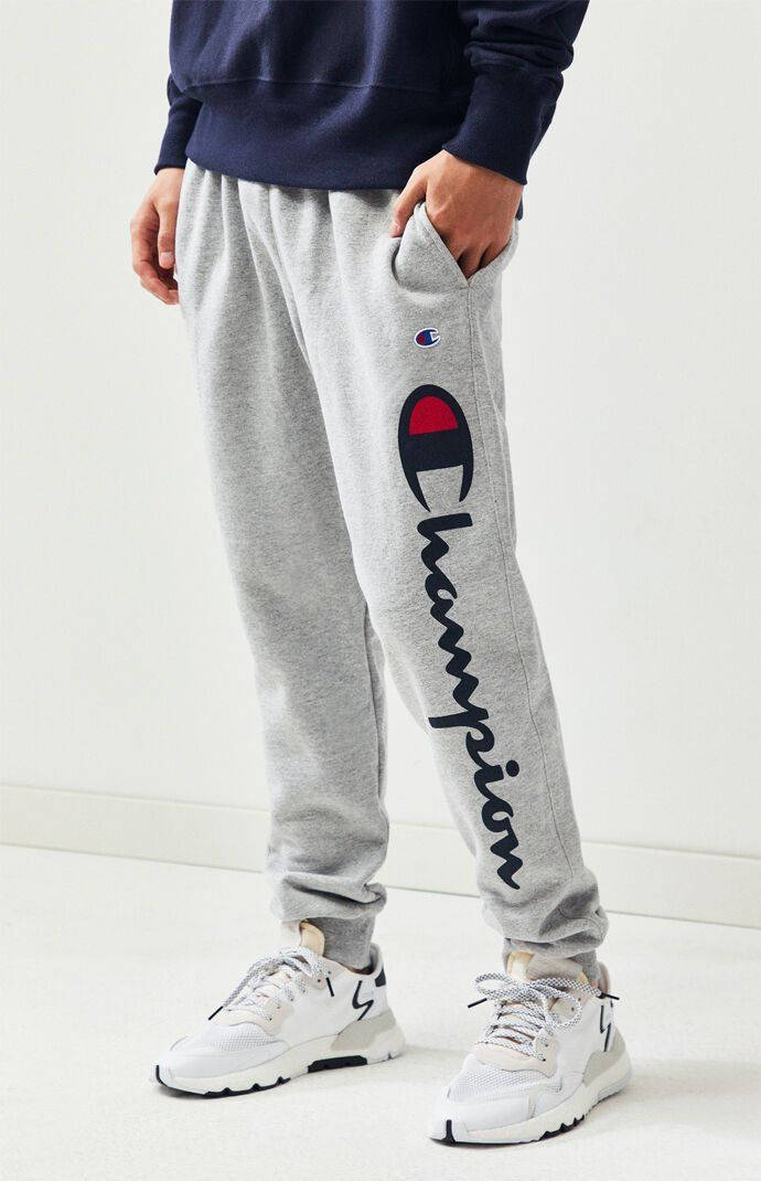 Champion Logo 桌布 壁紙 — 一個人穿著灰色 Champion 運動褲，褲管上有標誌，搭配白色運動鞋，背景為純白色。