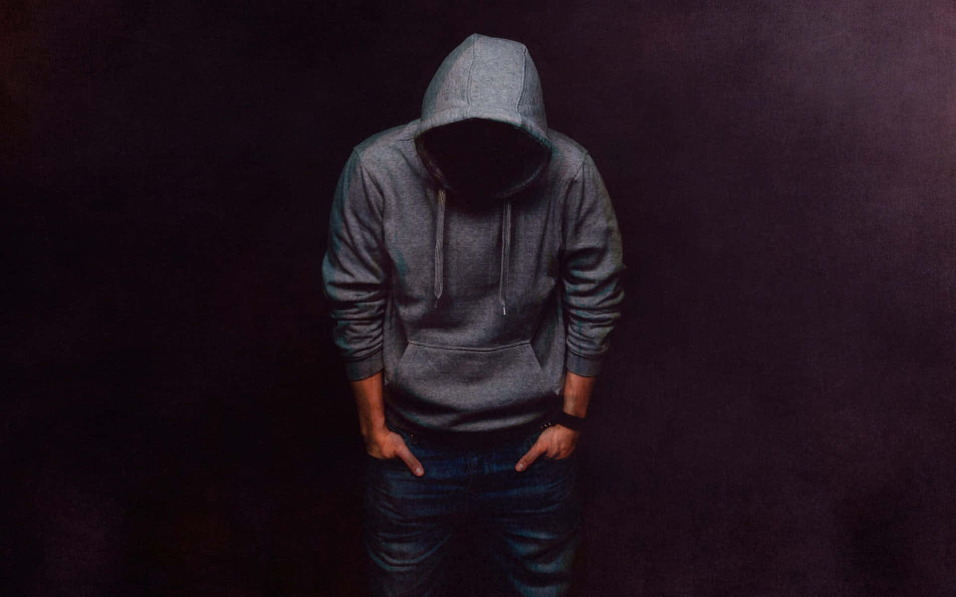Hoodie 桌布 壁紙 — 一個人穿著灰色連帽衫，低著頭，站在深紫色背景前，雙手插在牛仔褲口袋裡。