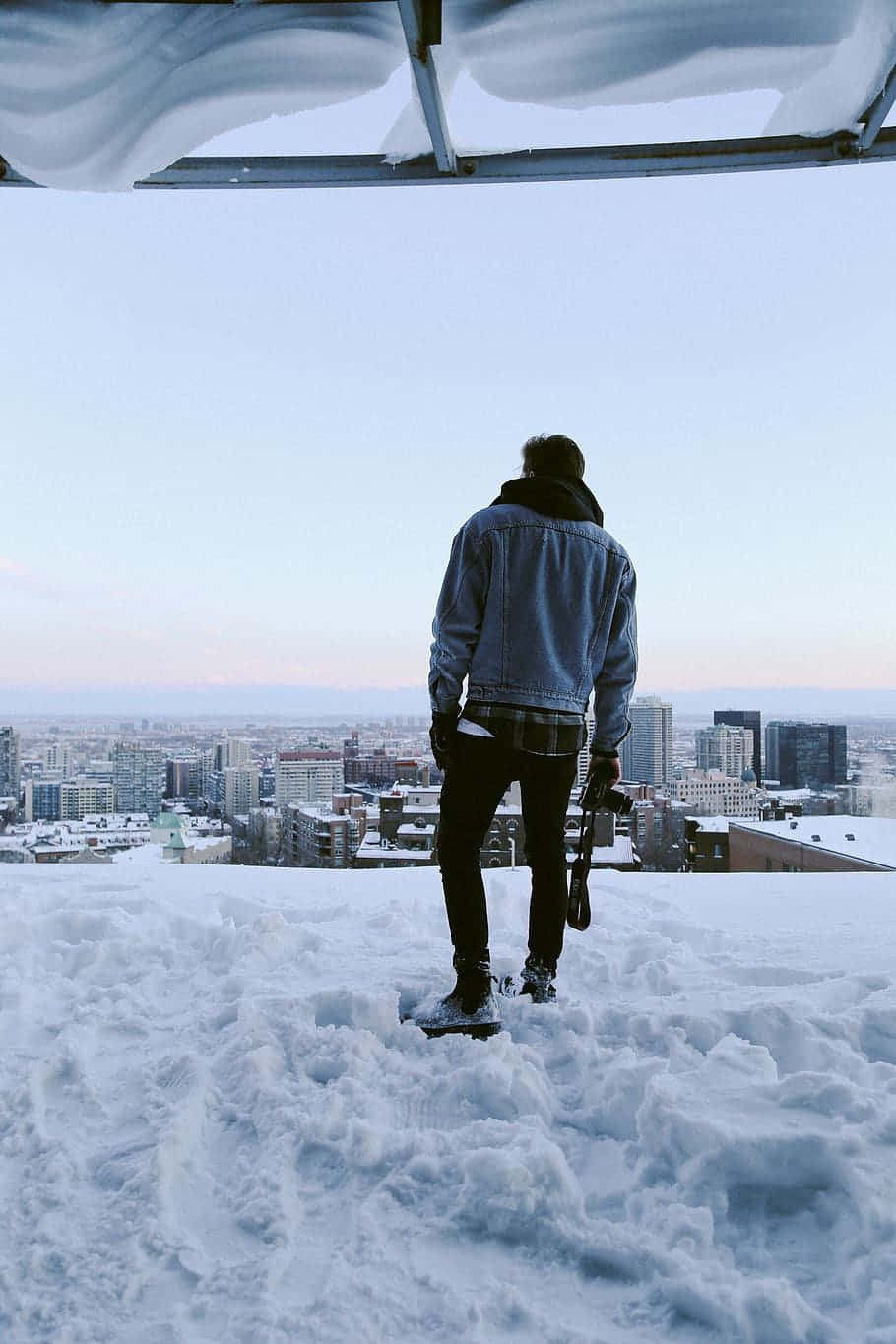 雪景 桌布 壁紙 — 一個人穿著牛仔外套，站在積雪的屋頂上，俯瞰黎明時分廣闊的冬季城市景觀。