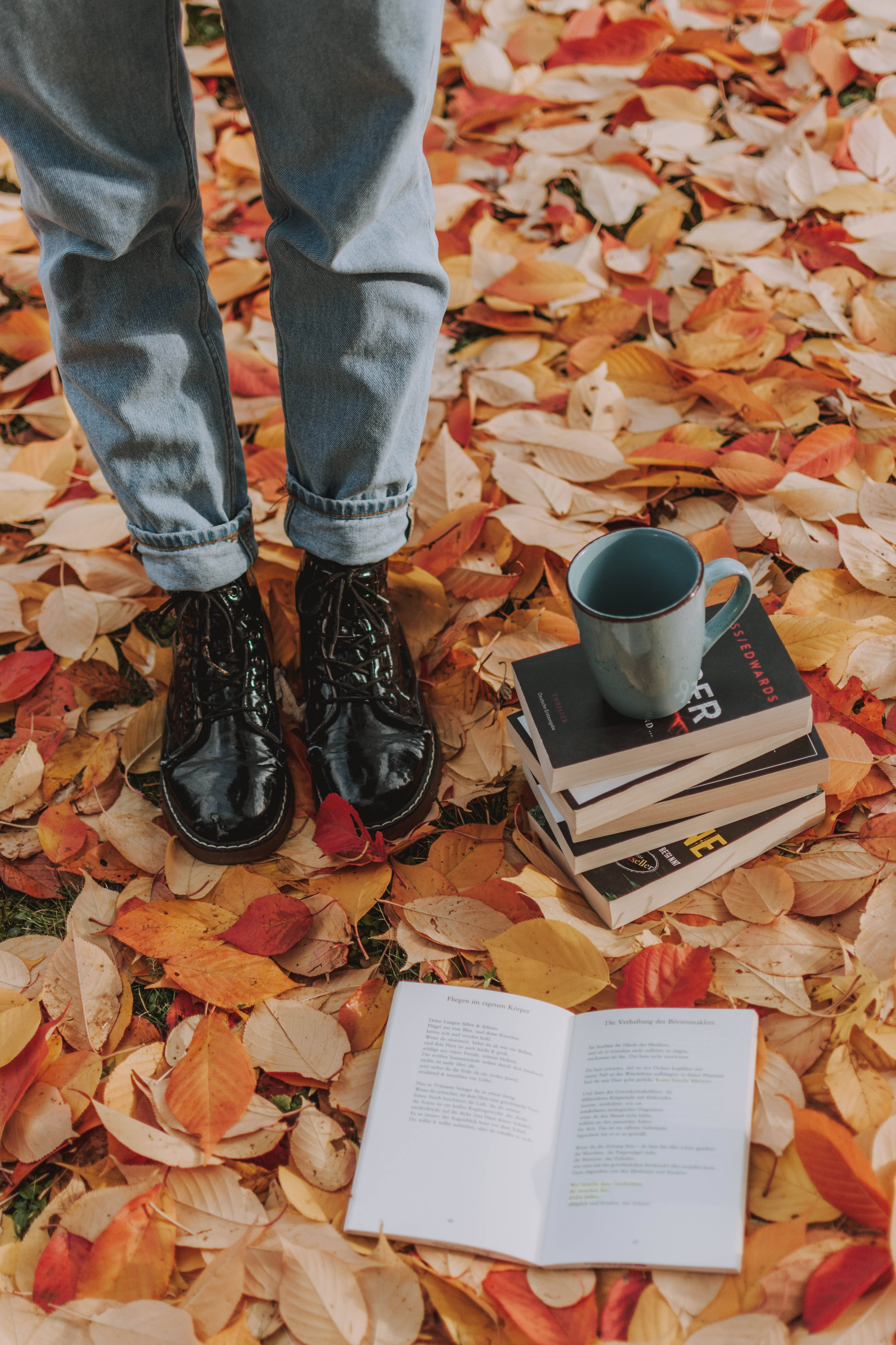 Fall Leaves 桌布 壁紙 — 一個人穿著牛仔褲和黑色靴子，站在鮮豔的秋葉中，有一本打開的書和一疊書，上面放著一個杯子。