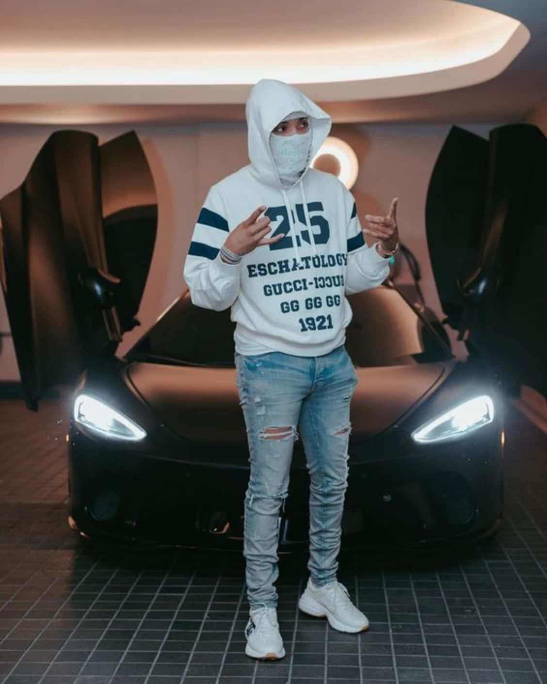 Herbo iPhone 桌布 壁紙 — 一個人穿著白色連帽衫和破洞牛仔褲，自信地站在一輛黑色跑車前，車門像海鷗翅膀一樣打開，在一個時尚的車庫裡。