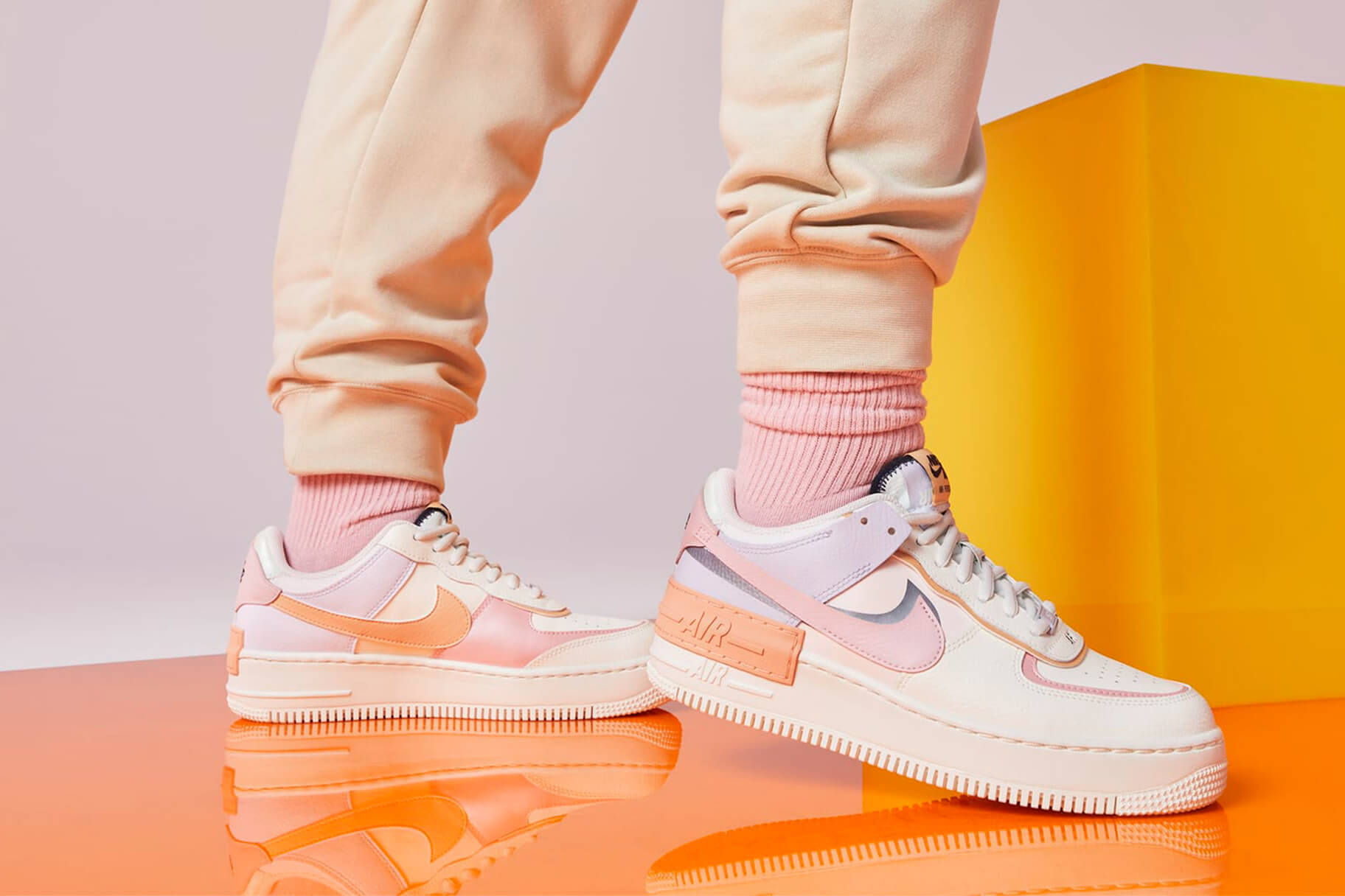 Nike Shoes 桌布 壁紙 — 一個人穿著粉彩粉紅和蜜桃色的Nike Air Force 1運動鞋，搭配相配的襪子和運動褲，站在反光的橙色表面上，背景是鮮明的黃色區塊和柔和的粉紅色。