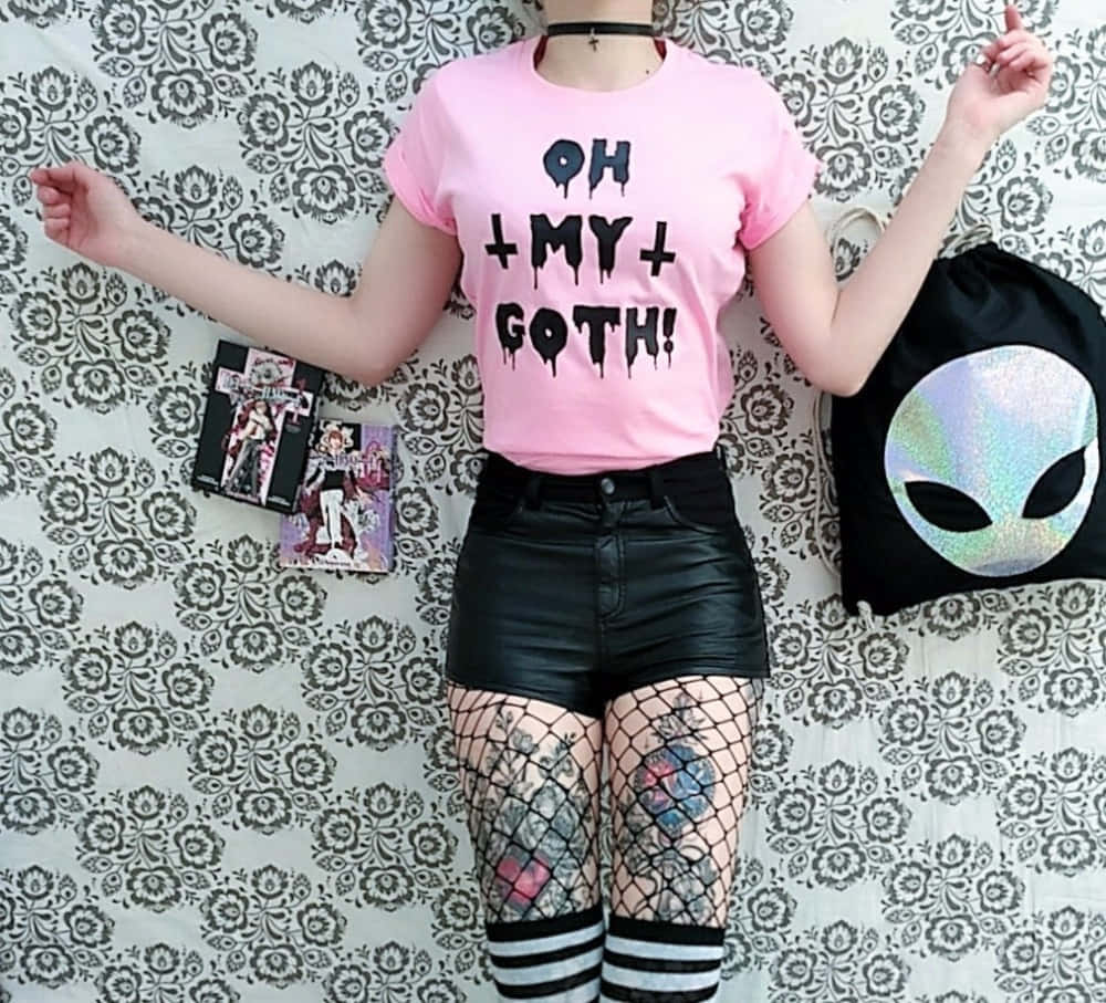 垃圾美學 桌布 壁紙 — 一個人穿著粉紅色「OH MY GOTH！」T恤和黑色皮革短褲，在花卉壁紙前擺姿勢，帶著外星人背包和漫畫書。