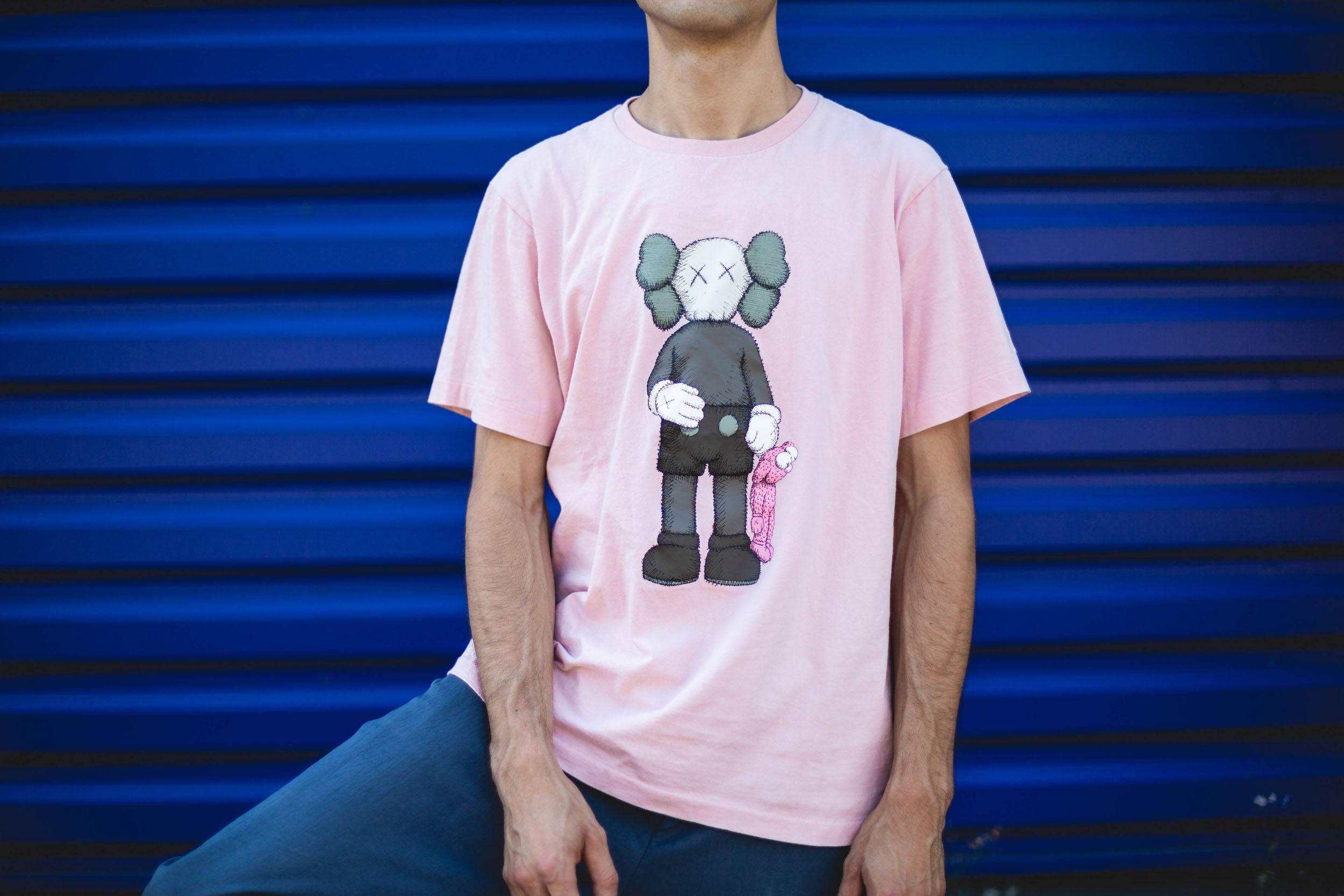 KAWS 桌布 壁紙 — 一個人穿著粉紅色T恤，上面有KAWS角色拿著粉紅色滑板，背景是充滿活力的藍色波紋金屬牆。