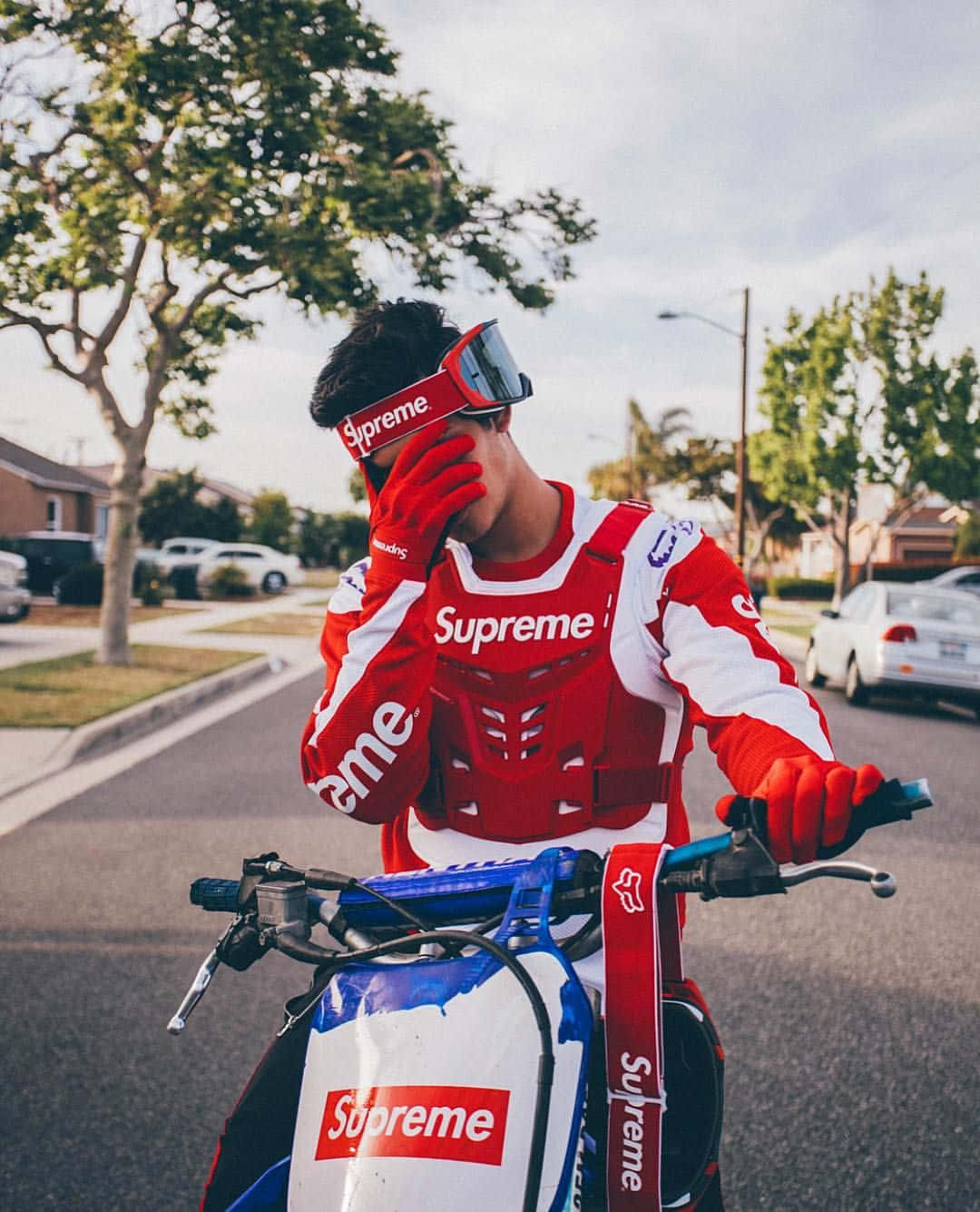 穿著紅色 Supreme 裝備的人，在陽光明媚的郊區街道上，在品牌滑板車上調整護目鏡。 — Valorant Logo 壁紙