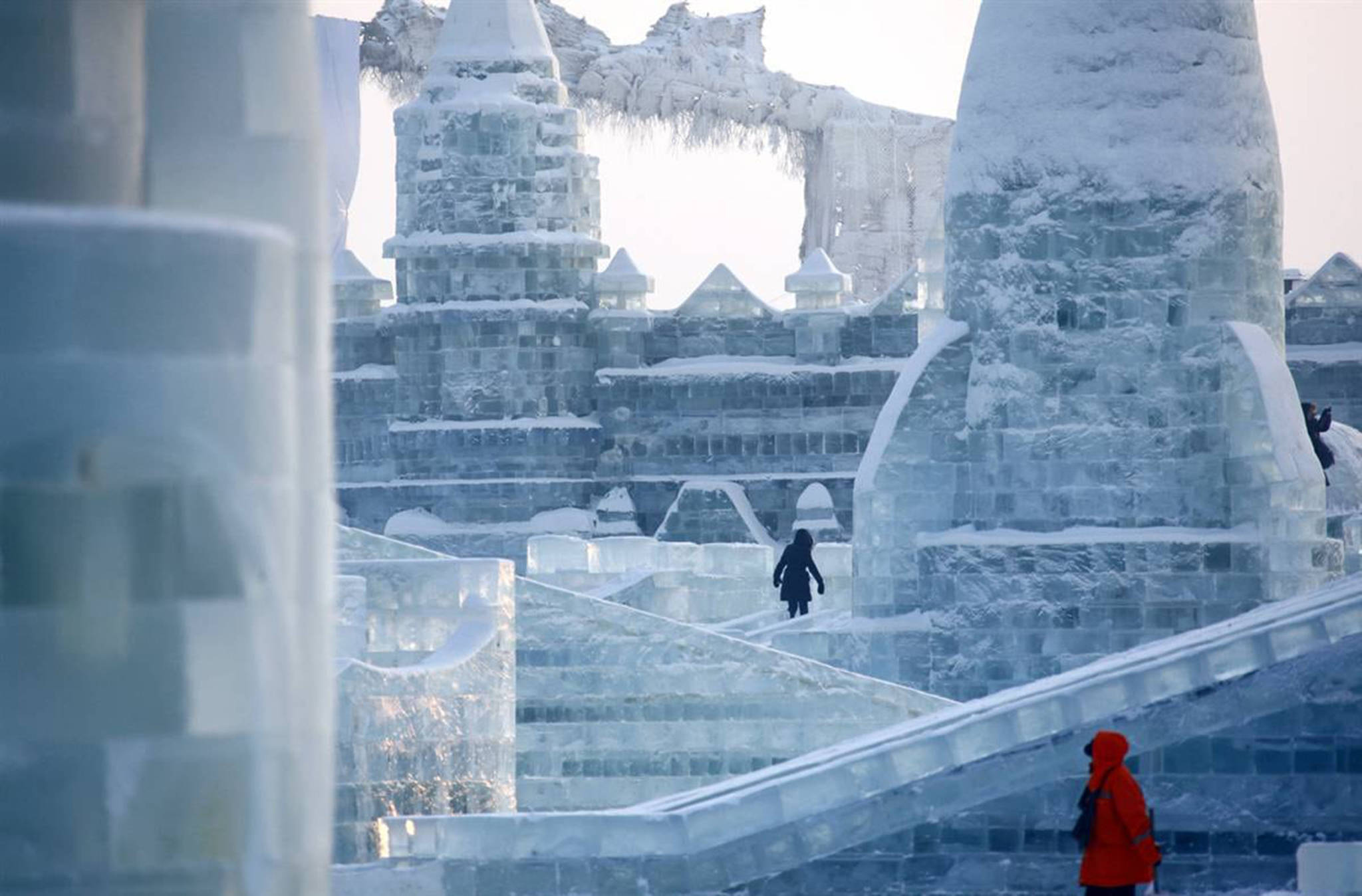 Frozen Castle 桌布 壁紙 — 一個人穿著紅色外套，在明亮的冬日天空下，走過一座由半透明藍色方塊組成的宏偉冰城堡。