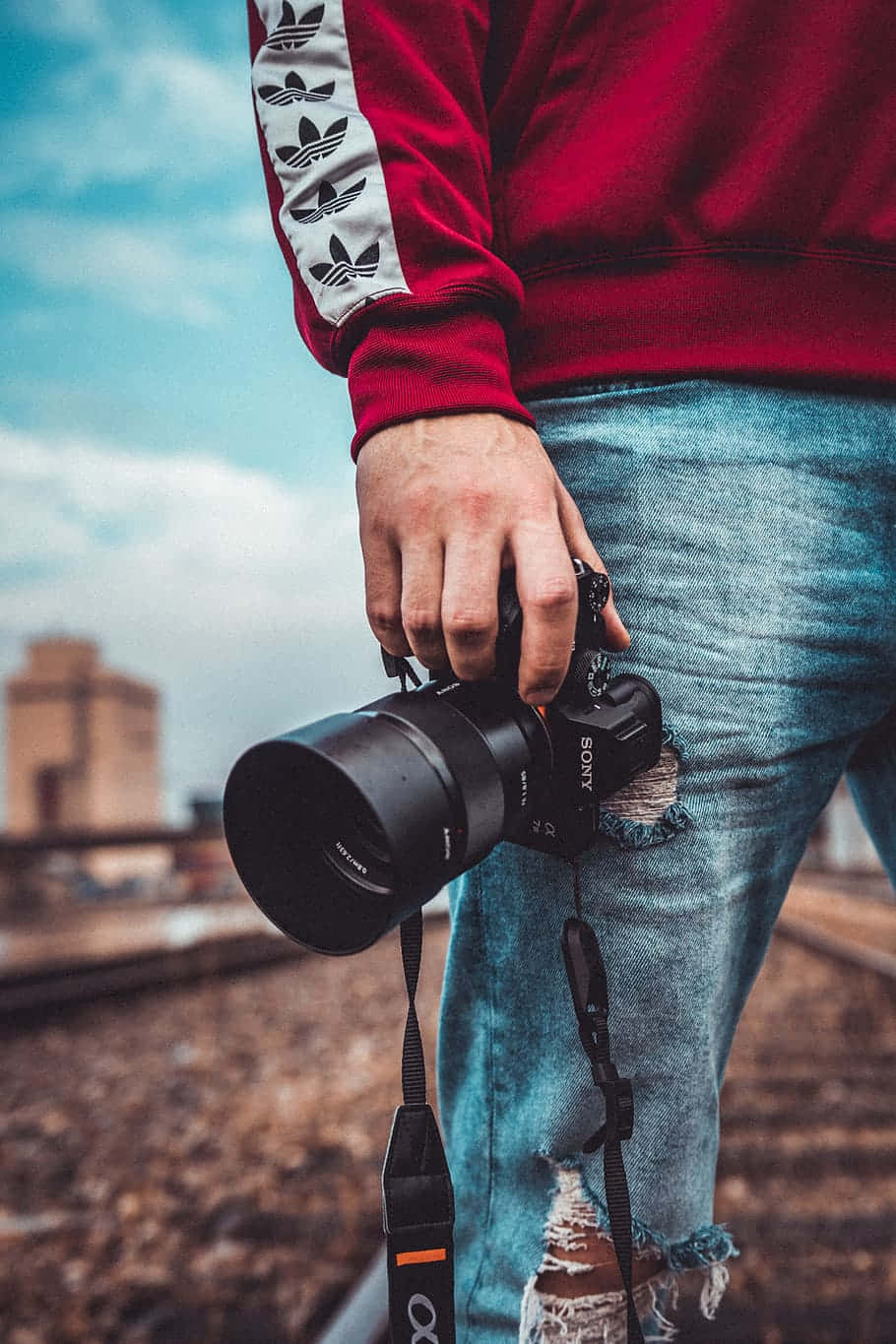 Dslr Camera 桌布 壁紙 — 一個人穿著紅色連帽衫和破洞牛仔褲，在屋頂上拿著一部黑色Sony相機，背景是陰天下模糊的城市建築。
