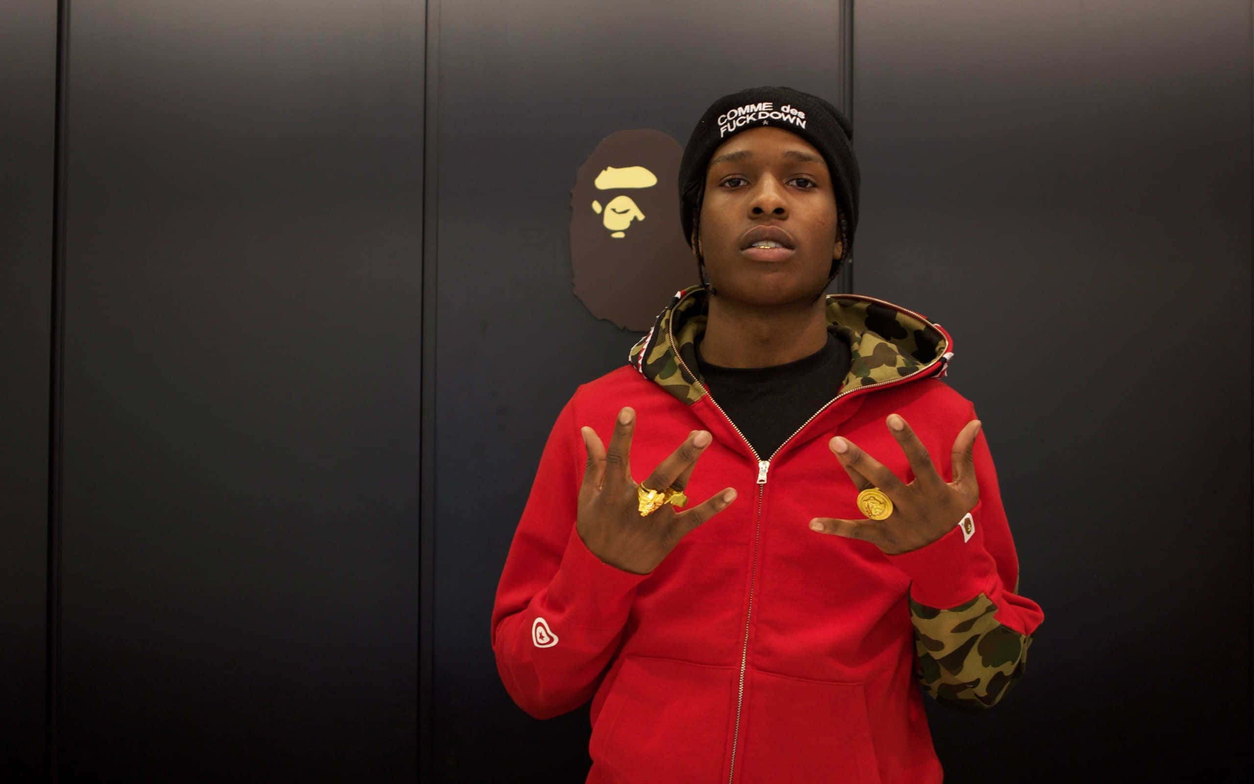 ASAP Rocky 桌布 壁紙 — 一個人穿著紅色連帽衫和黑色毛線帽，站在帶有BAPE標誌的深色牆壁前，舉起雙手展示金戒指。