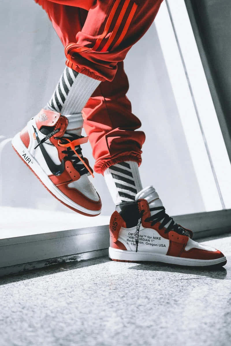 Off White Jordan 1 桌布 壁紙 — 一個人穿著紅色運動褲和帶黑色鞋帶的醒目紅白運動鞋，站在明亮窗戶附近的混凝土邊緣上。
