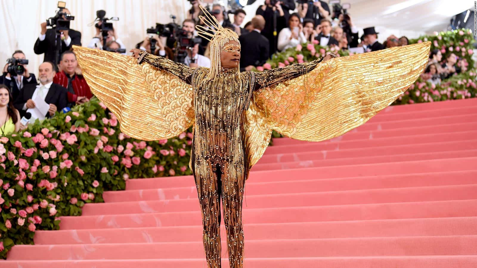Met Gala 桌布 壁紙 — 一個人穿著耀眼金色天使服裝，展開翅膀，在攝影師和粉紅玫瑰的簇擁下，沿著鋪有粉紅地毯的樓梯走下來。