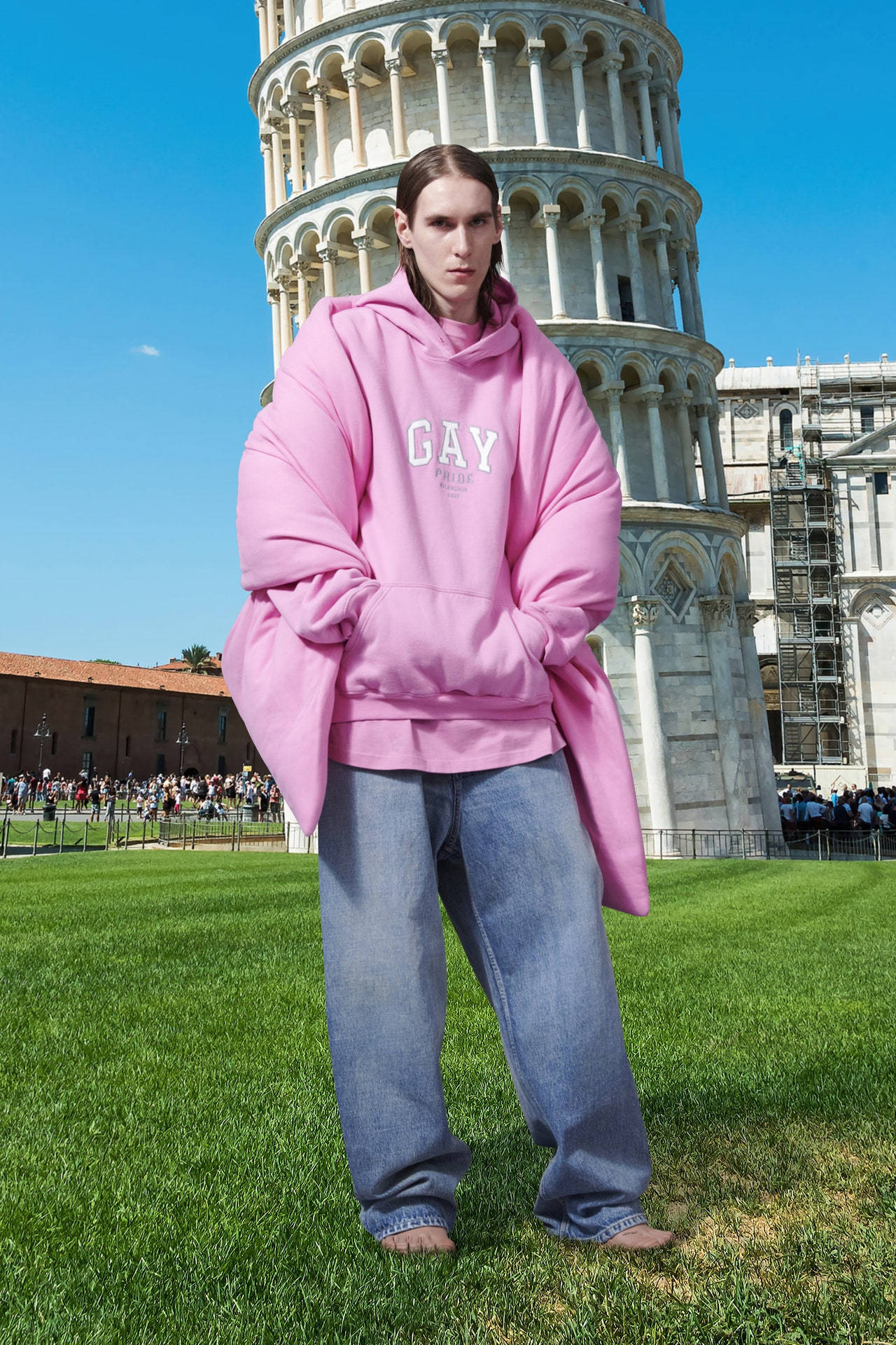 Balenciaga 桌布 壁紙 — 一個人穿著超大粉紅色連帽衫，赤腳站在比薩斜塔前的草地上，天空湛藍。