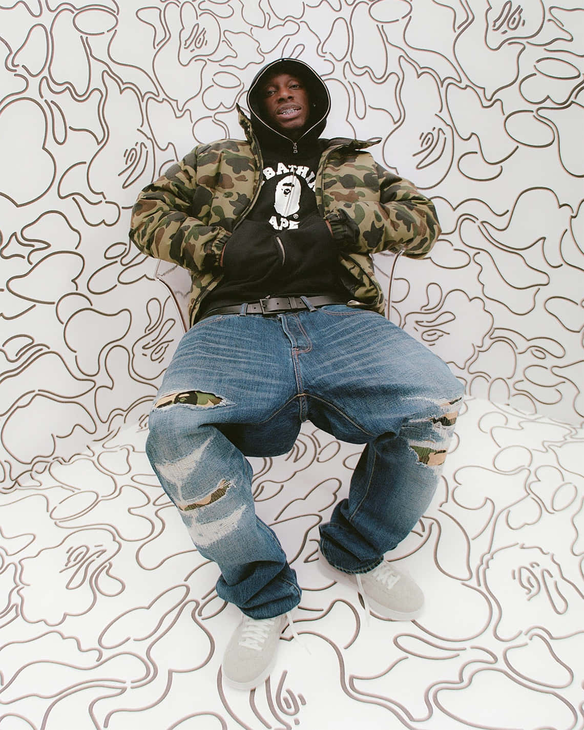 Bape 動態 桌布 壁紙 — 一個人穿著迷彩外套和破洞牛仔褲，自信地坐在白色牆壁前，牆上有棕色抽象線條藝術。
