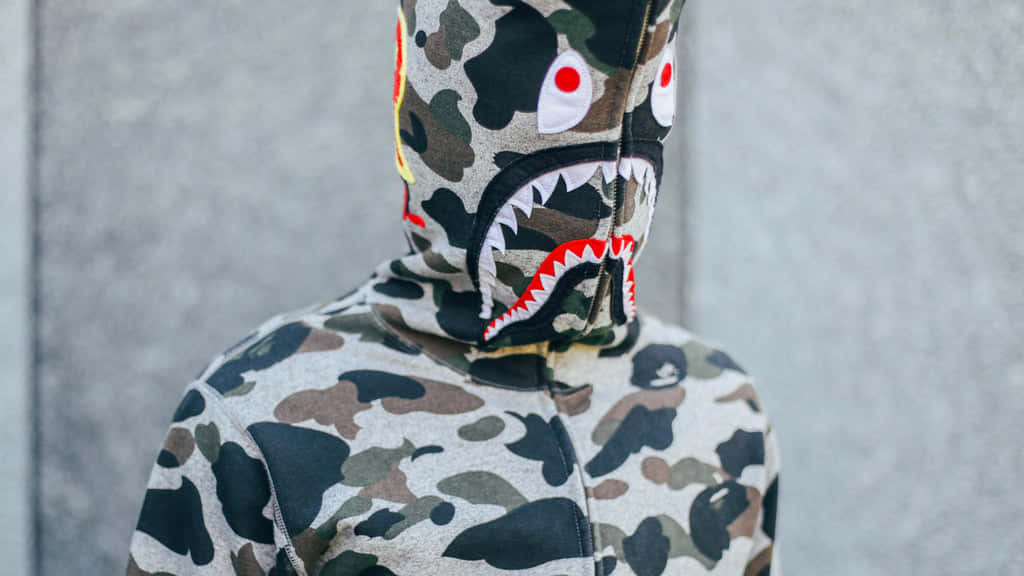 Bape 動態 桌布 壁紙 — 一個人穿著迷彩連帽衫，帽子上有鯊魚臉設計，背景是灰色混凝土牆。