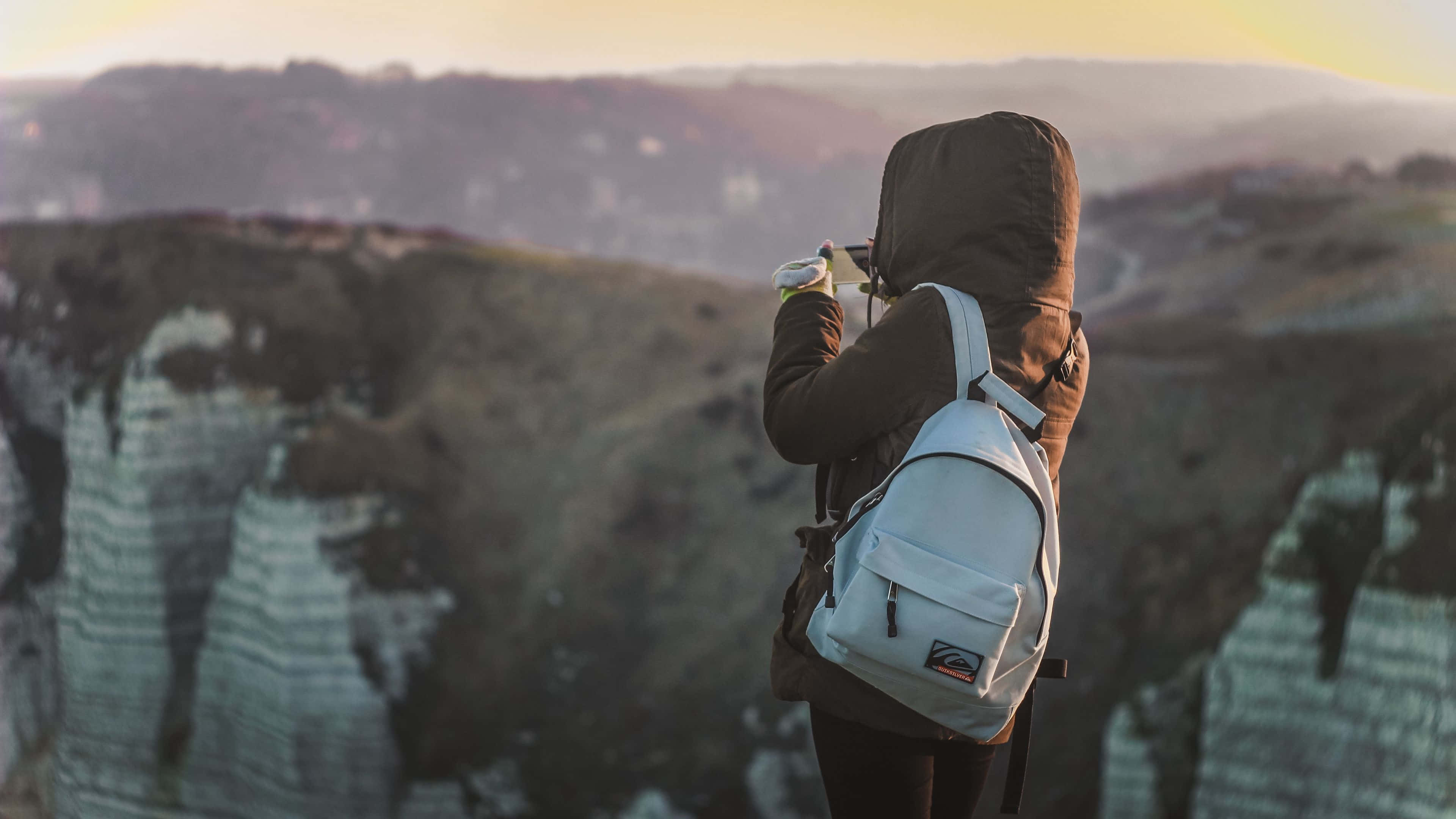 Traveler 桌布 壁紙 — 一個人穿著連帽外套和白色背包，在日落時站在懸崖邊，用相機捕捉廣闊的風景。