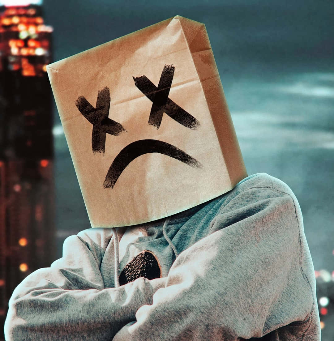 Marshmello 動態圖 桌布 壁紙 — 一個人穿著連帽衫，頭上戴著一個棕色的紙袋，上面畫著悲傷的臉，背景是夜晚模糊的城市天際線。
