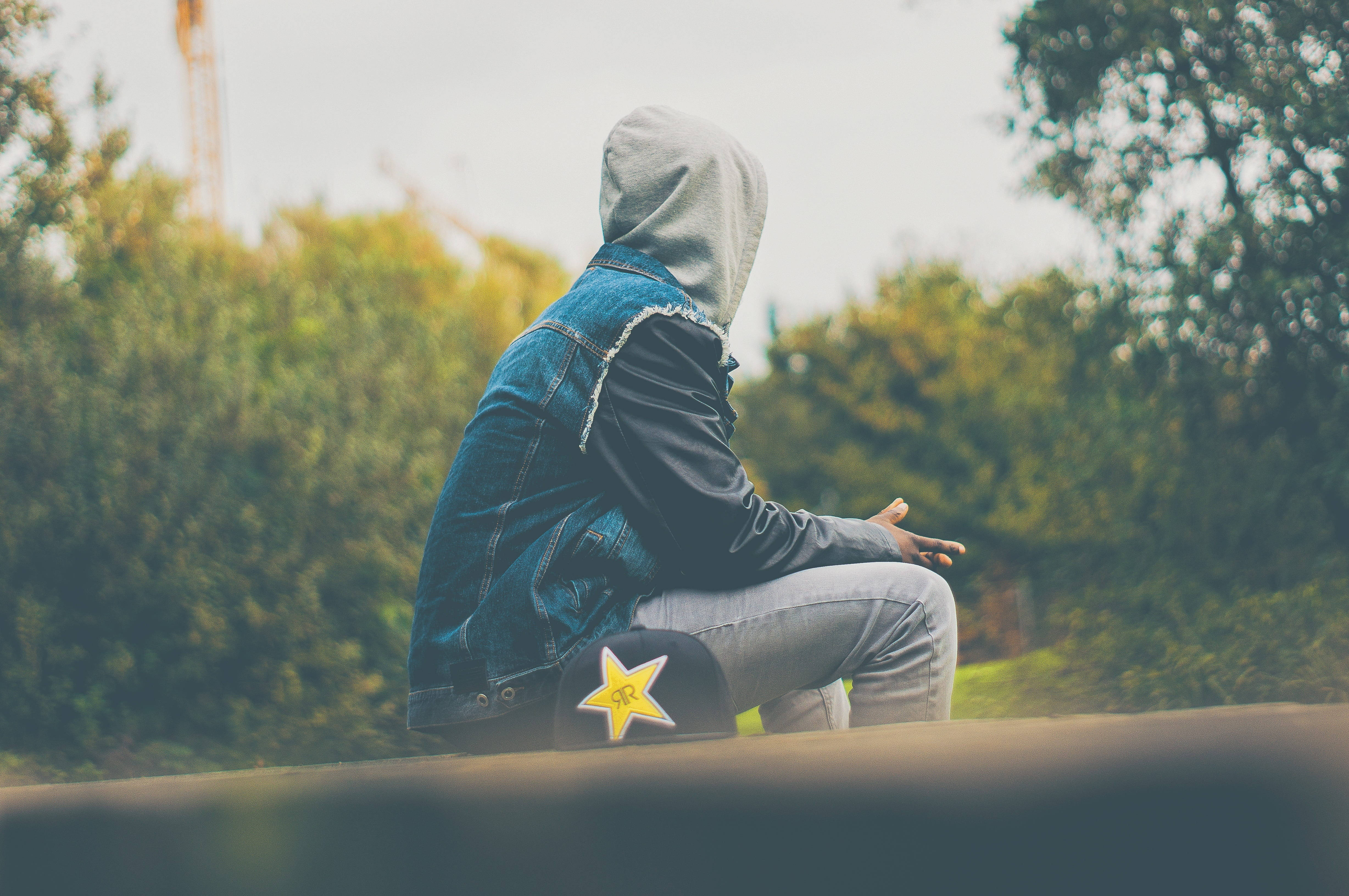 Alone Boy 桌布 壁紙 — 一個人穿著連帽衫和牛仔外套，獨自坐在一個俯瞰綠樹的壁架上，天空多雲。