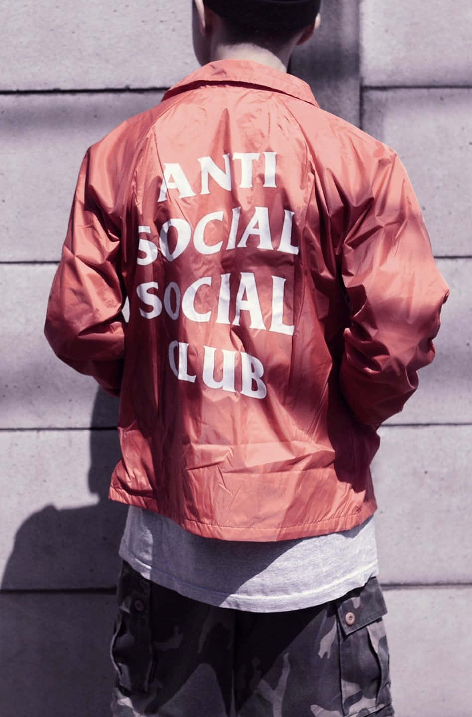 Anti Social Club iPhone 桌布 壁紙 — 一個人穿著鏽色 'ANTI SOCIAL SOCIAL CLUB' 外套，背對鏡頭，在明亮的陽光下靠著混凝土牆站立。