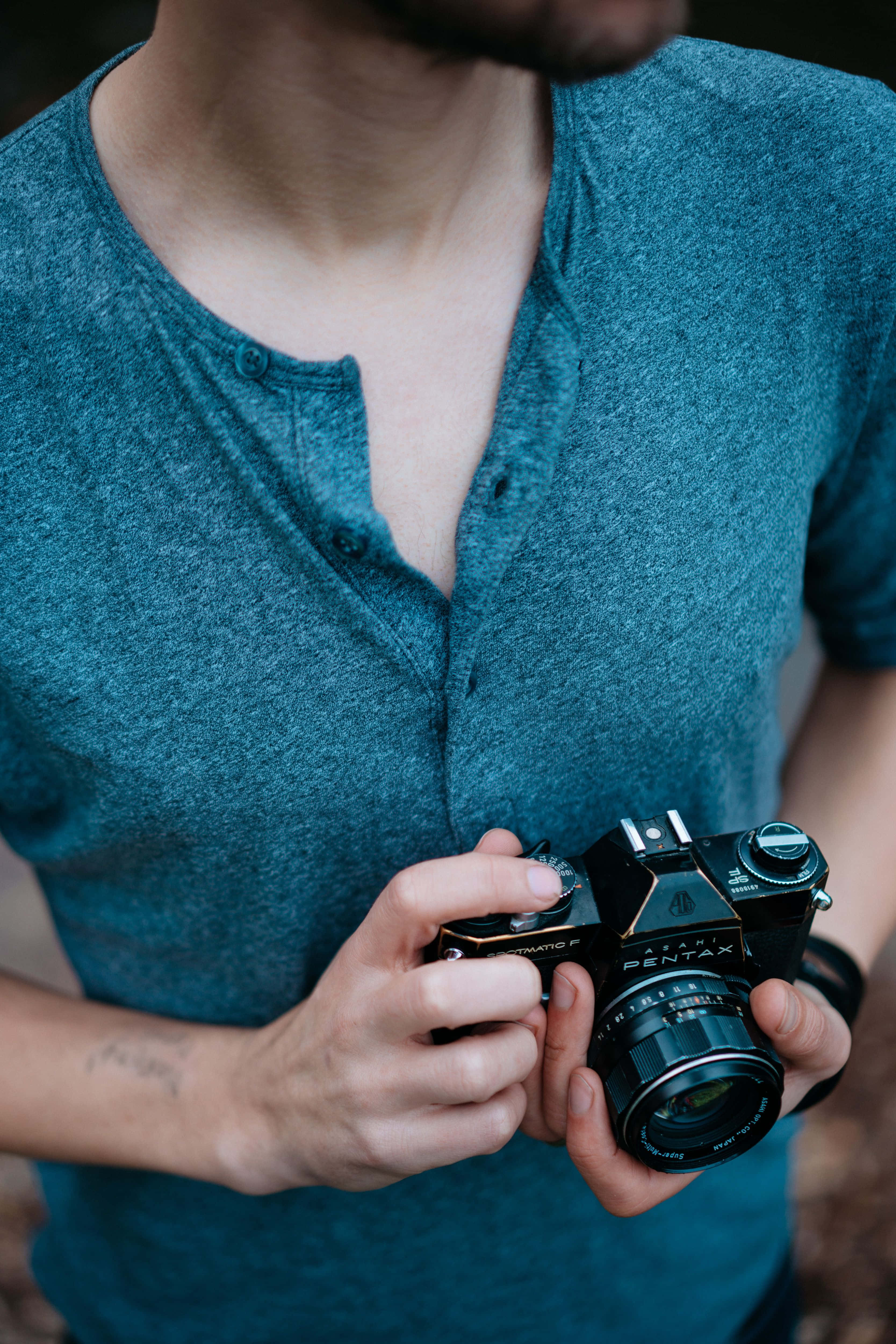 Holding Camera 桌布 壁紙 — 一個人穿著青綠色亨利衫，雙手拿著一部黑色賓得底片相機，背景是模糊的自然景色。