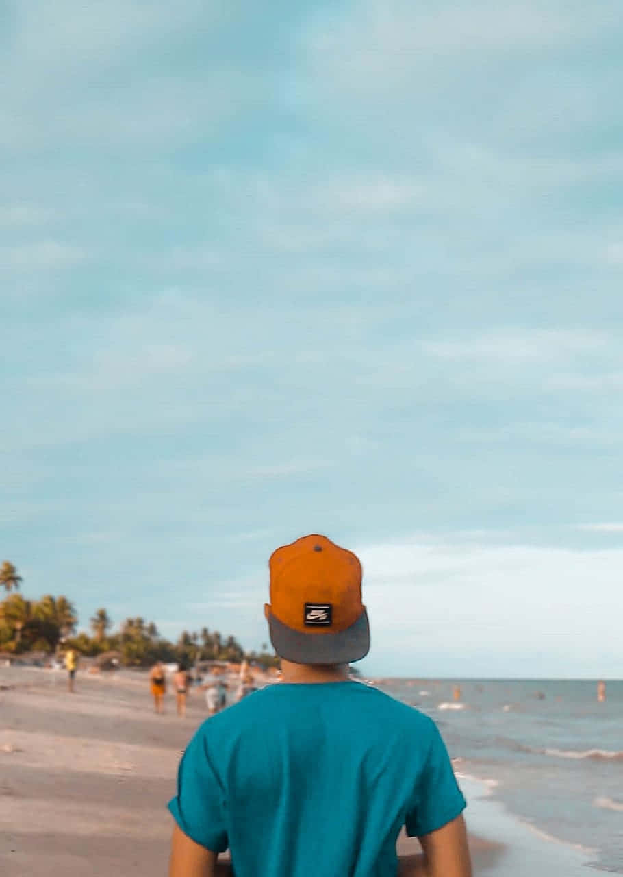 Traveler 桌布 壁紙 — 一個人穿著青綠色襯衫和橙色帽子，在柔和的藍天下沿著沙灘漫步，伴隨著溫柔的海浪。