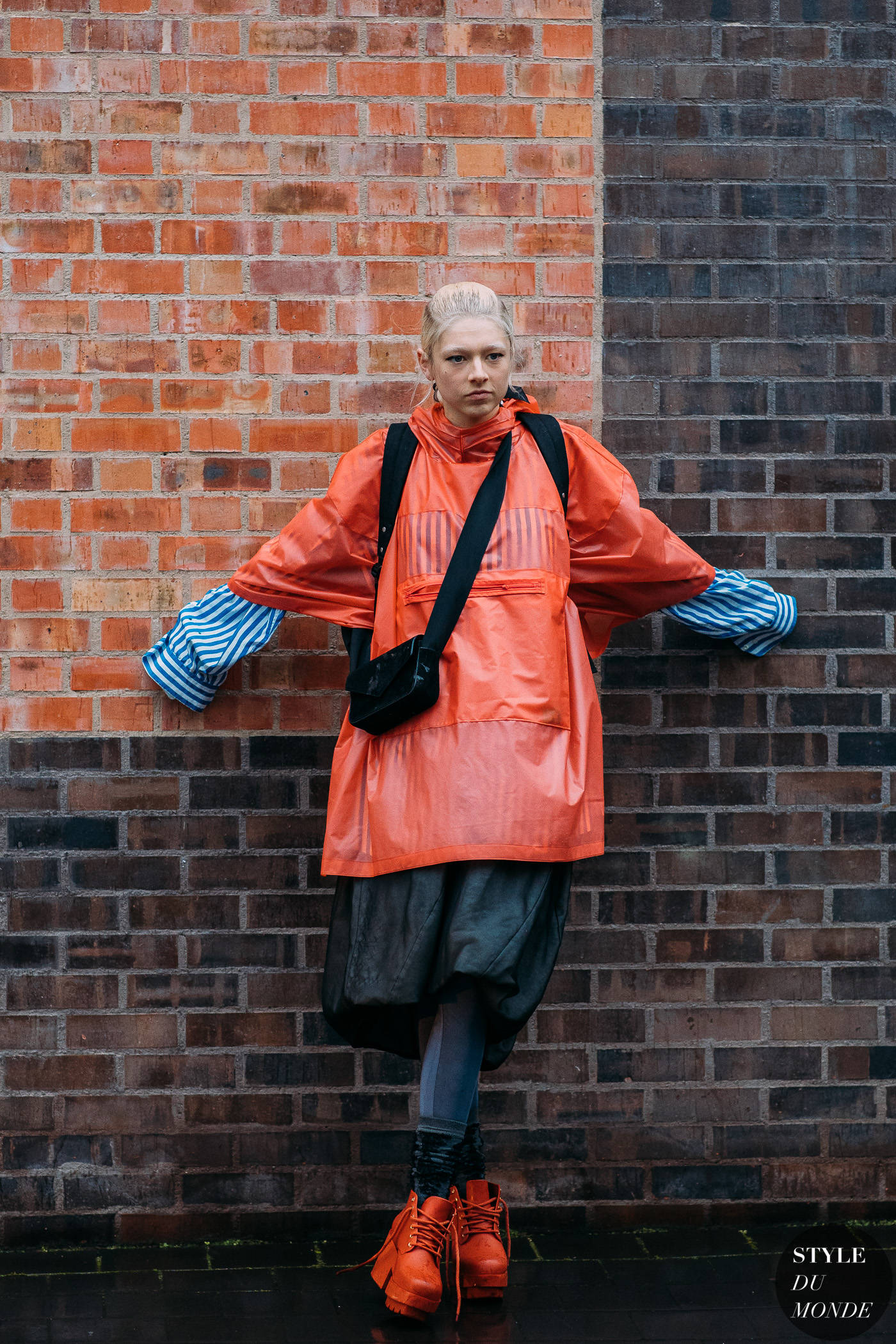 Hunter Schafer 桌布 壁紙 — 一個人穿著鮮橙色雨衣和條紋袖子，自信地站在風化的磚牆前。