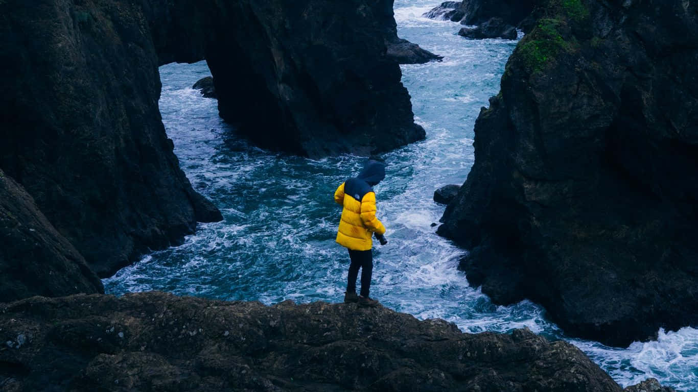 Traveler 桌布 壁紙 — 一個人穿著鮮黃色外套，站在岩石懸崖上，俯瞰著在黑暗懸崖之間撞擊的洶湧藍色海水。