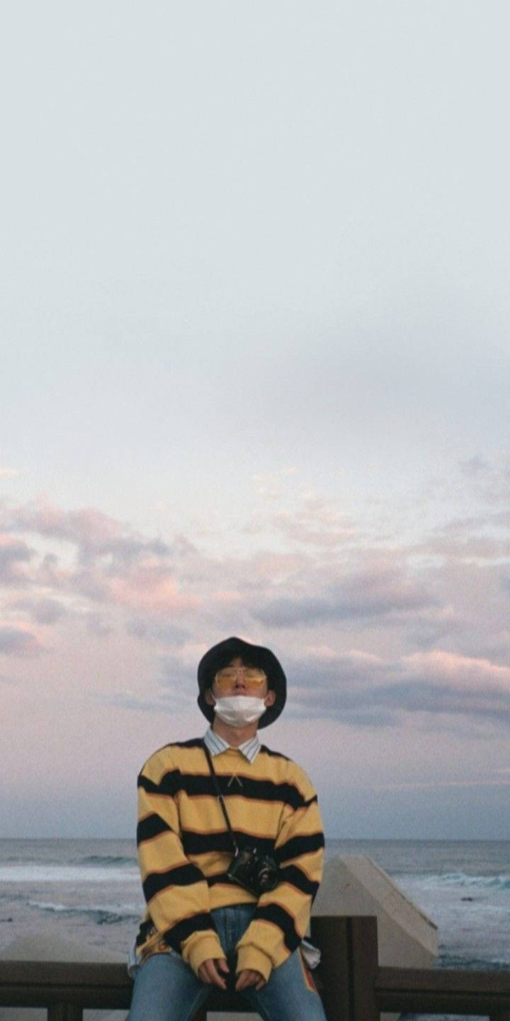 kim hanbin 桌布 壁紙 — 一個人穿著黃色條紋毛衣和口罩坐在木碼頭上，背景是柔和的粉紅色和灰色黃昏天空。