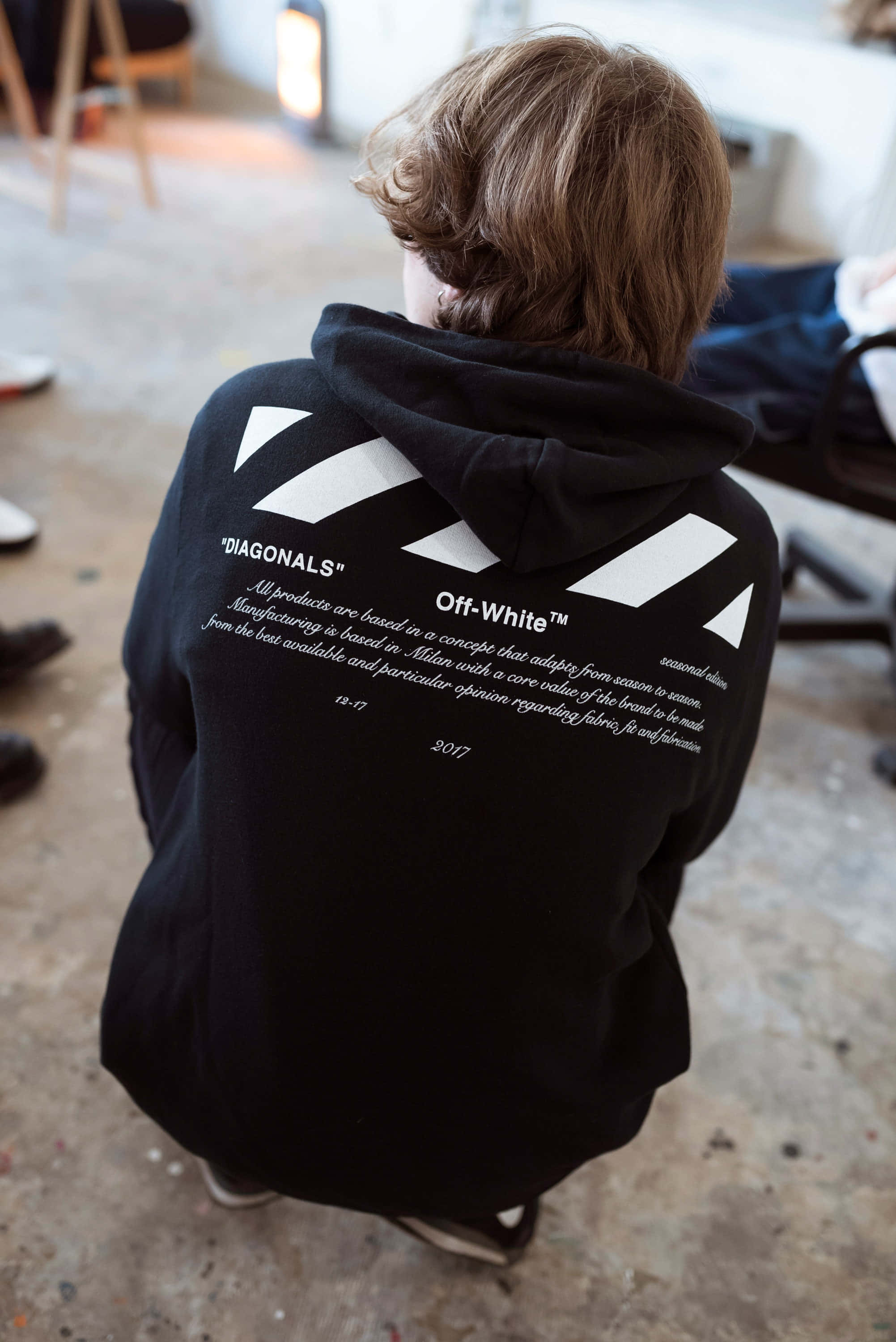 Off White 桌布 壁紙 — 一個人穿著黑色 Off-White 連帽衫，帶有白色斜紋和文字，從背後看到在雜亂的室內空間。