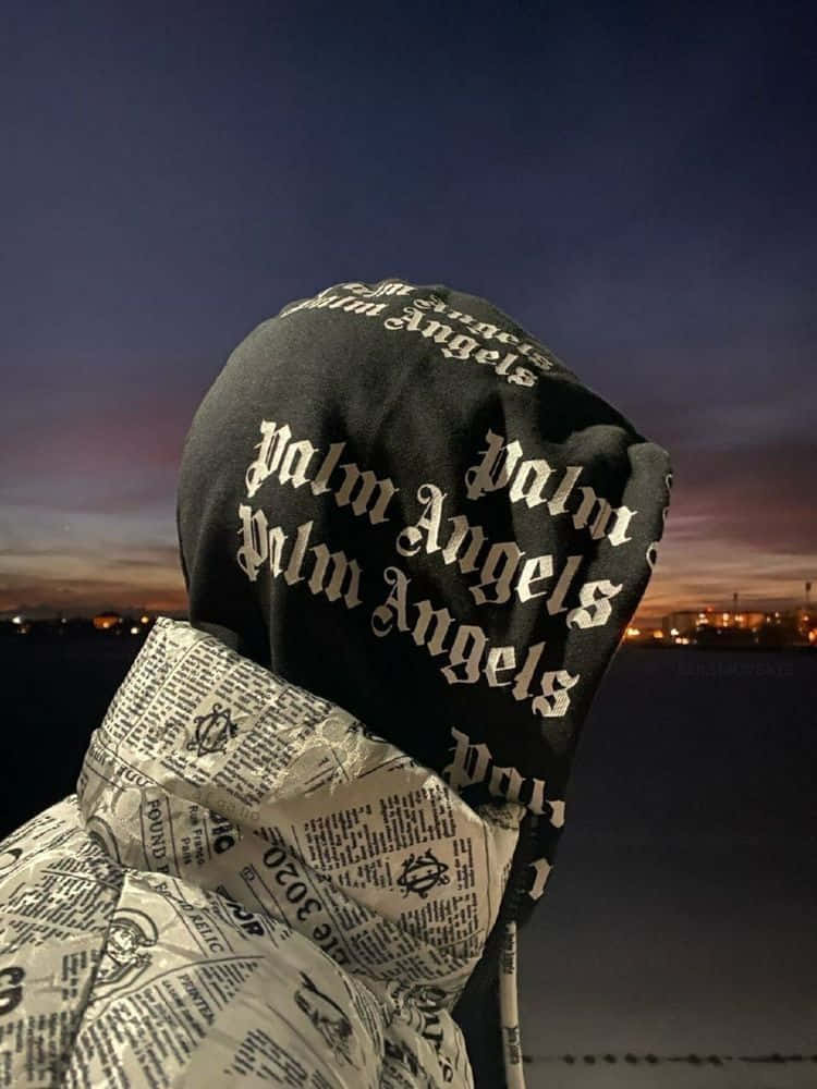 Hoodie 桌布 壁紙 — 一個人穿著黑色 'Palm Angels' 連帽衛衣和報紙印花外套，背對鏡頭，背景是暮色天空和遠處的城市燈光。