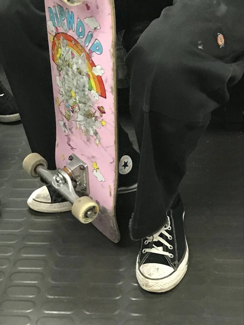 滑板 桌布 壁紙 — 一個人穿著黑色Converse運動鞋的腳部特寫，旁邊有一個粉紅色Ripndip滑板，輪子已磨損，放在有紋理的灰色地板上。