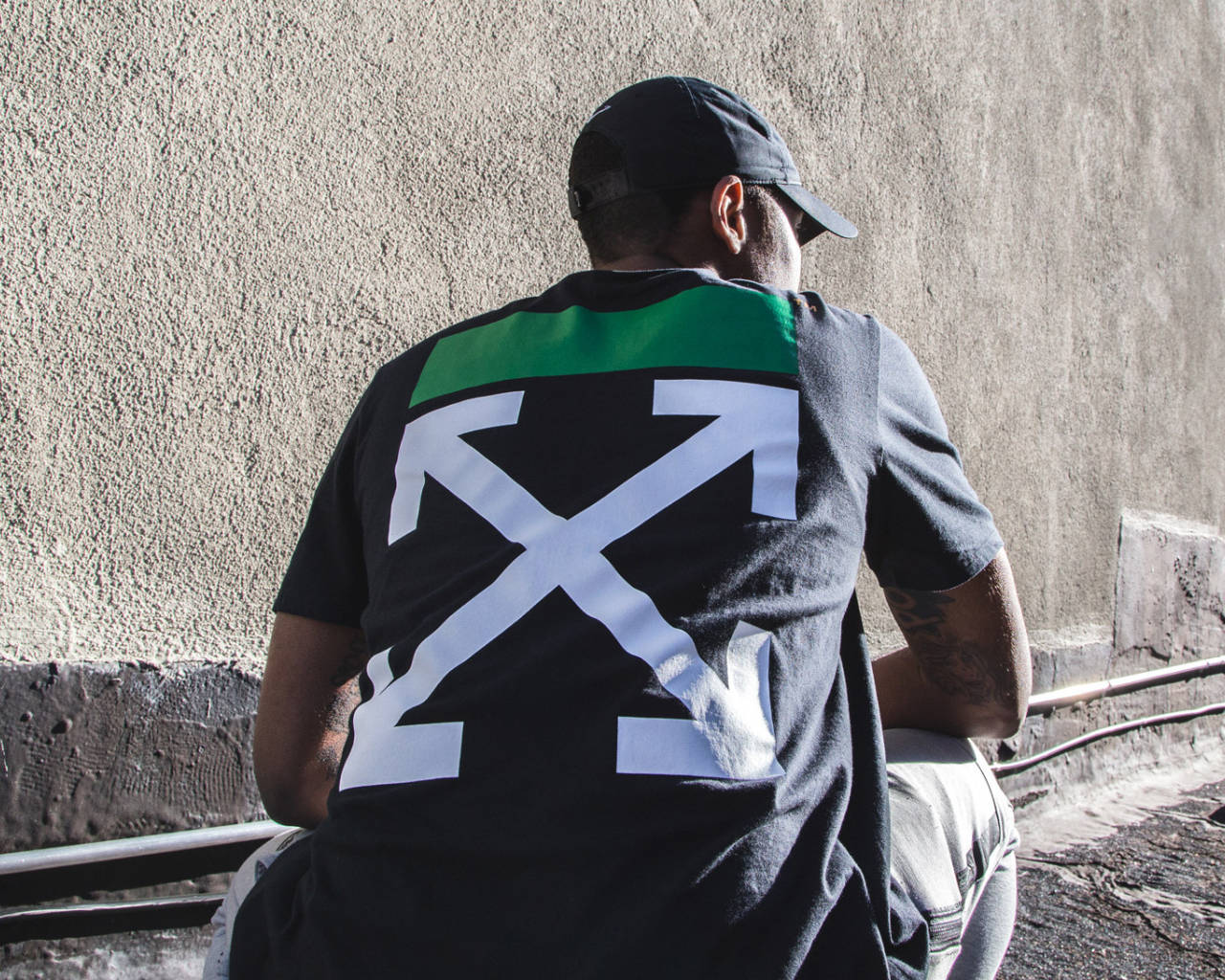 Off White Logo 桌布 壁紙 — 一個人穿著黑色T恤，上面有大型白色箭頭標誌和綠色條紋，靠著紋理混凝土牆坐著。