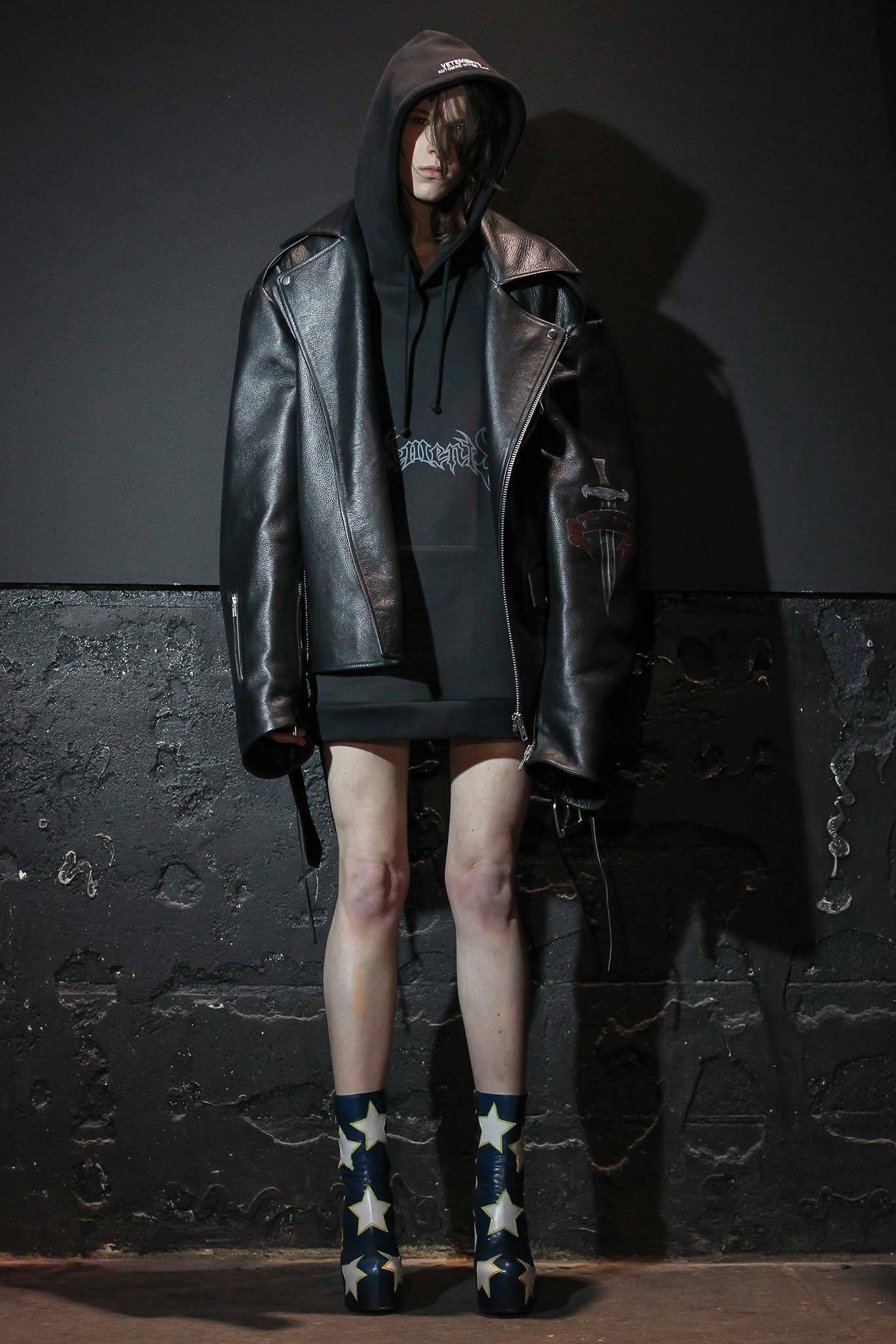 Vetements 桌布 壁紙 — 一個人穿著黑色皮夾克和星形圖案靴子，在戲劇性燈光下靠著深色紋理牆壁站立。