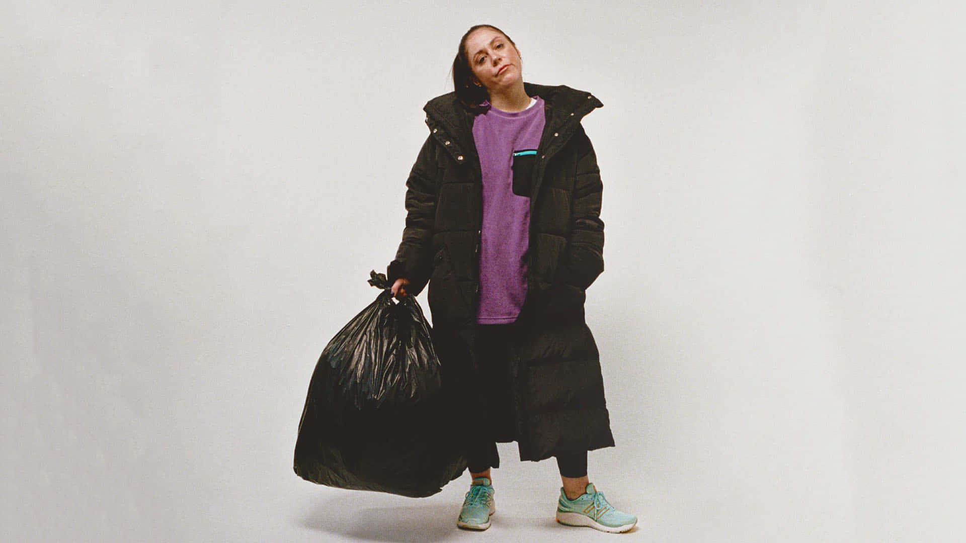 New Balance 桌布 壁紙 — 一個人穿著黑色羽絨外套和紫色襯衫，在純白色背景前拿著一個大的黑色垃圾袋。