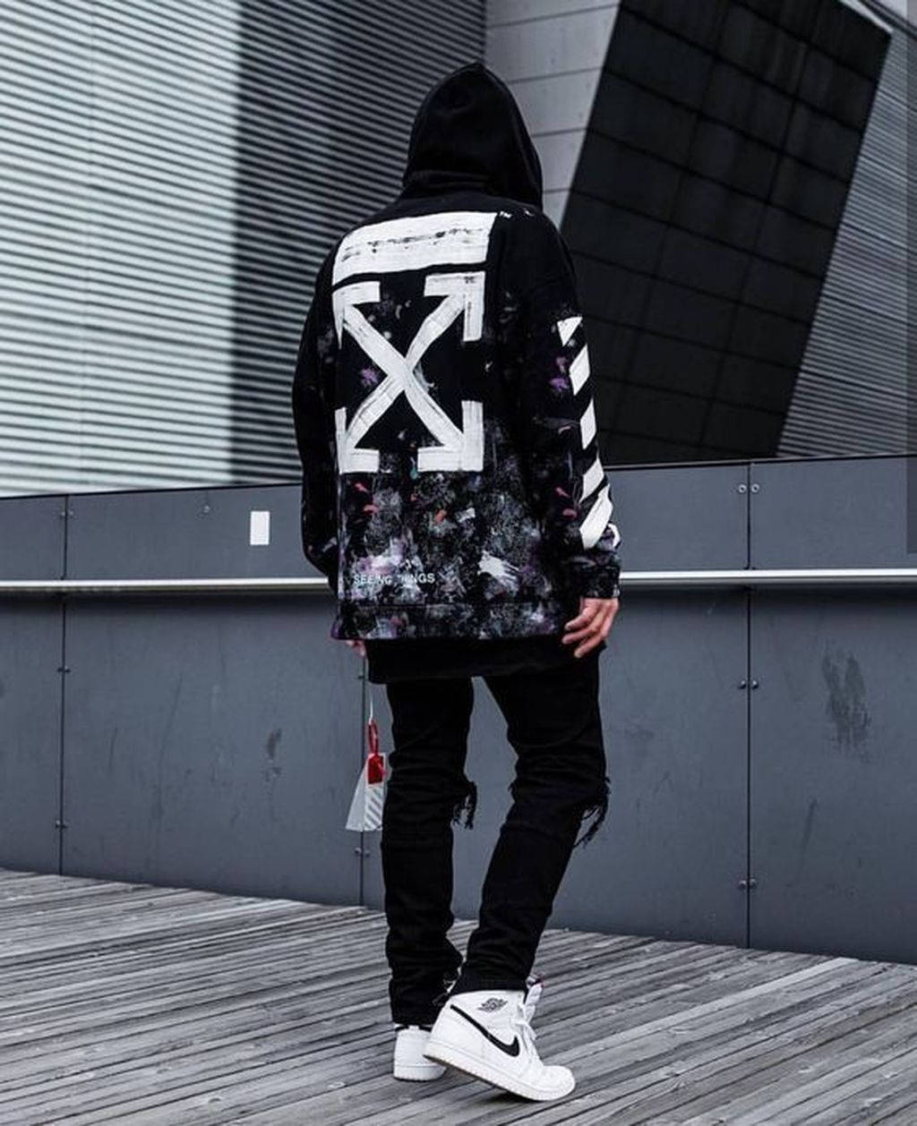 Off White Logo 桌布 壁紙 — 一個人穿著黑色連帽衫，上面有一個大的白色箭頭標誌，站在木甲板上，背景是現代灰色建築外牆。