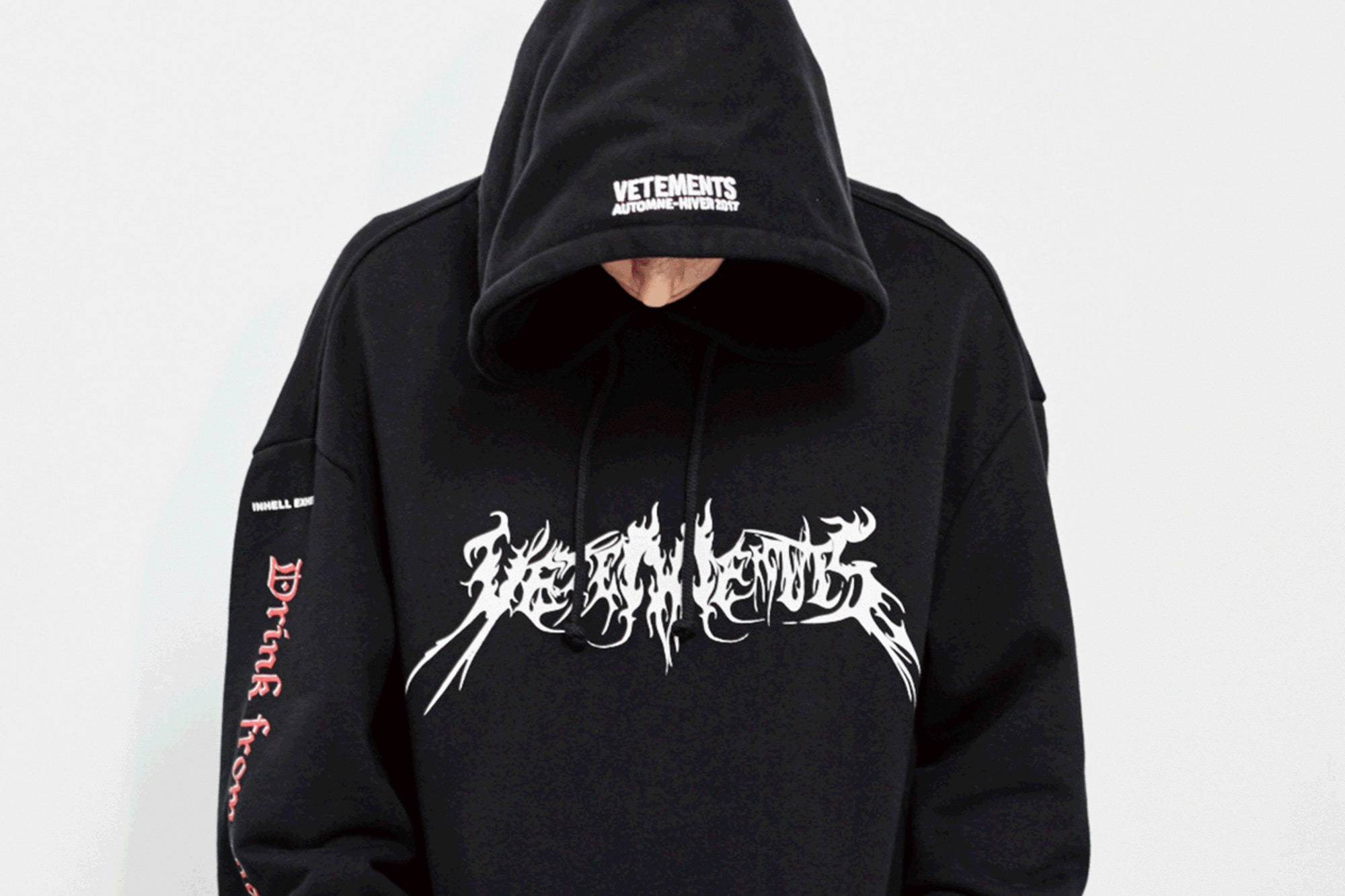 Vetements 桌布 壁紙 — 一個人穿著黑色連帽衫，上面有白色塗鴉文字和紅色袖子字母，臉藏在帽子下，背景是純白色。