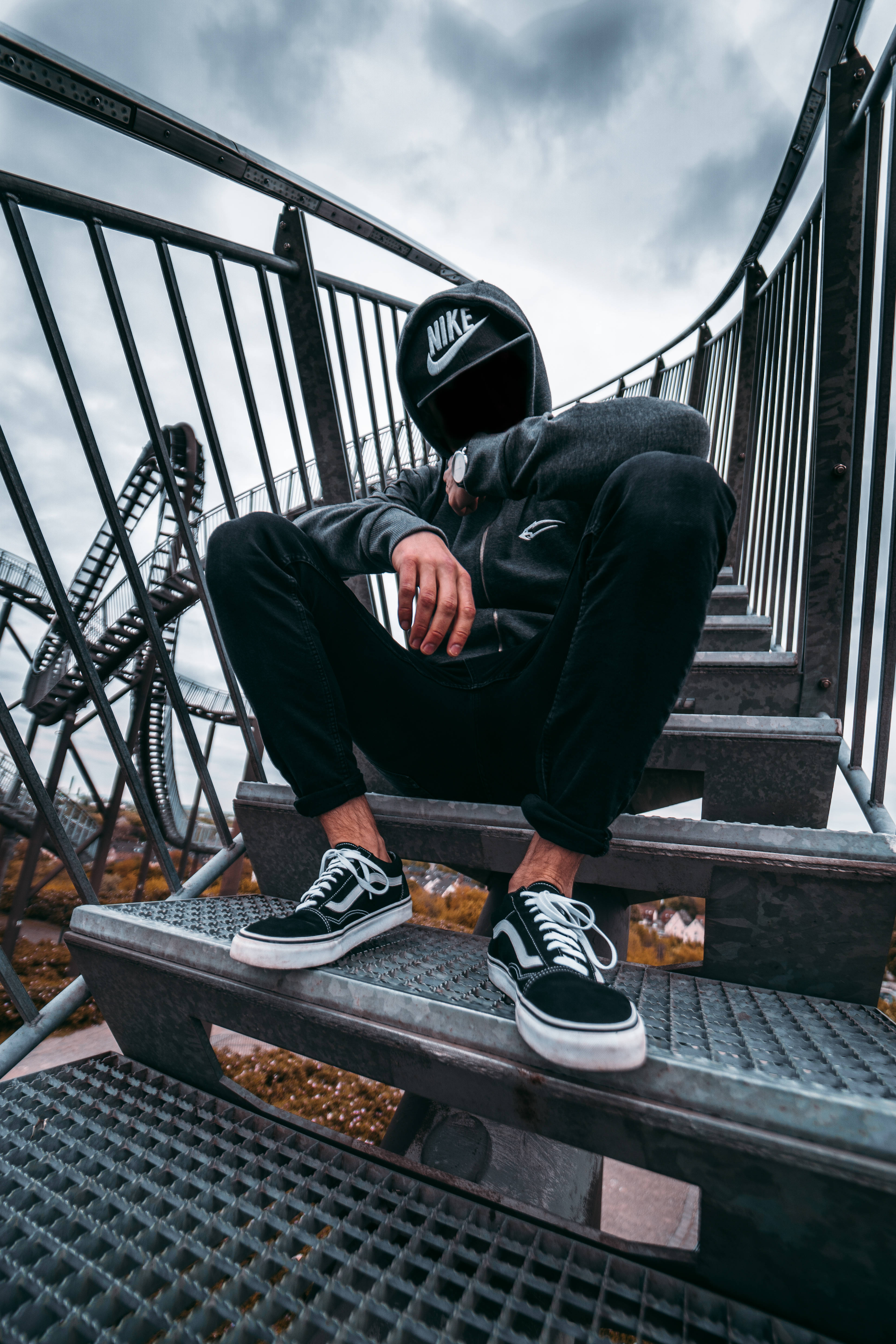 4K nike 桌布 壁紙 — 一個人穿著黑色連帽衫和Vans運動鞋坐在金屬樓梯上，背景是雲天下的一個雲霄飛車。