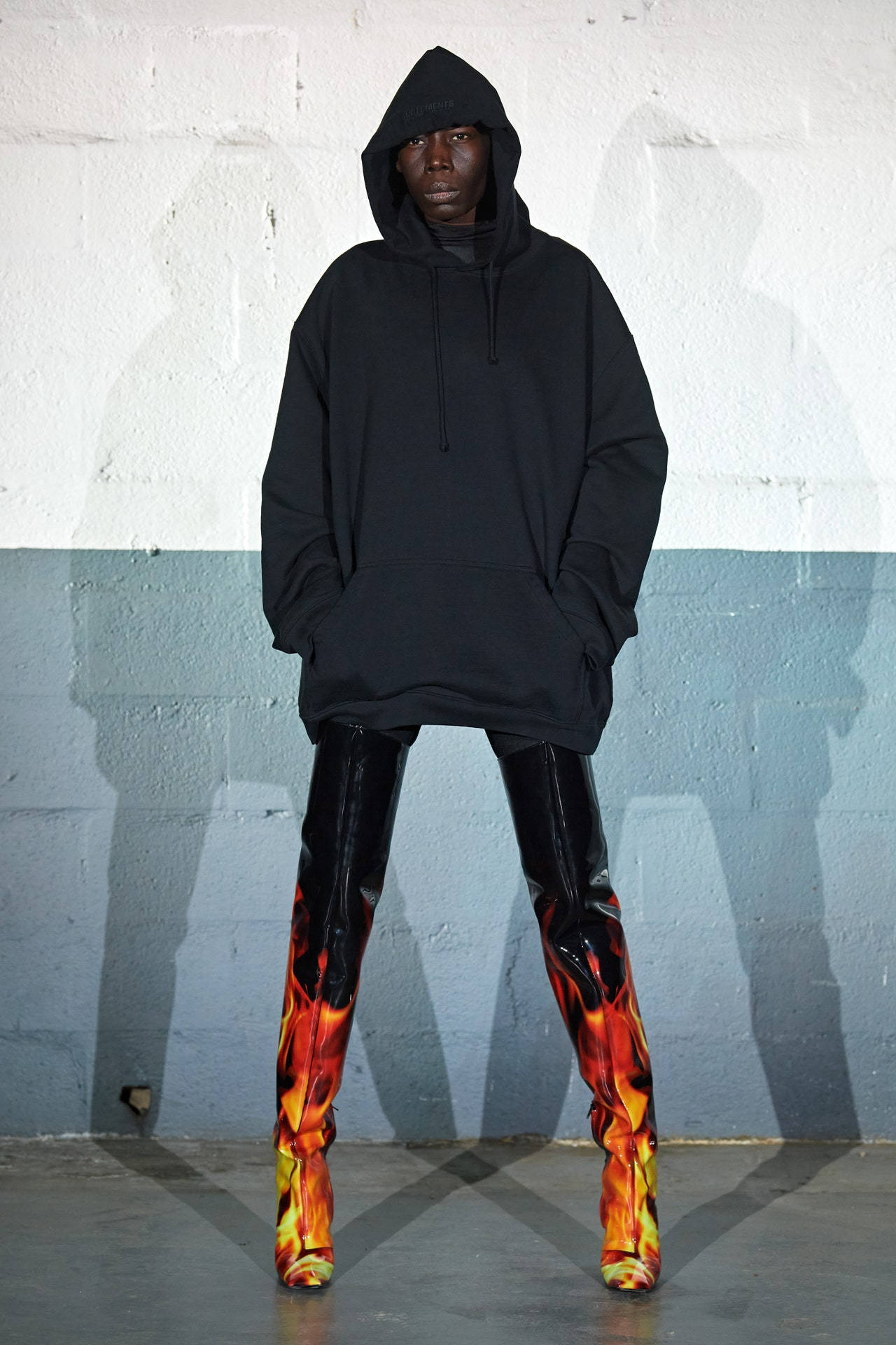 Vetements 桌布 壁紙 — 一個人穿著黑色連帽衫和火焰圖案靴子，站在雙色牆前，投射出戲劇性的陰影。