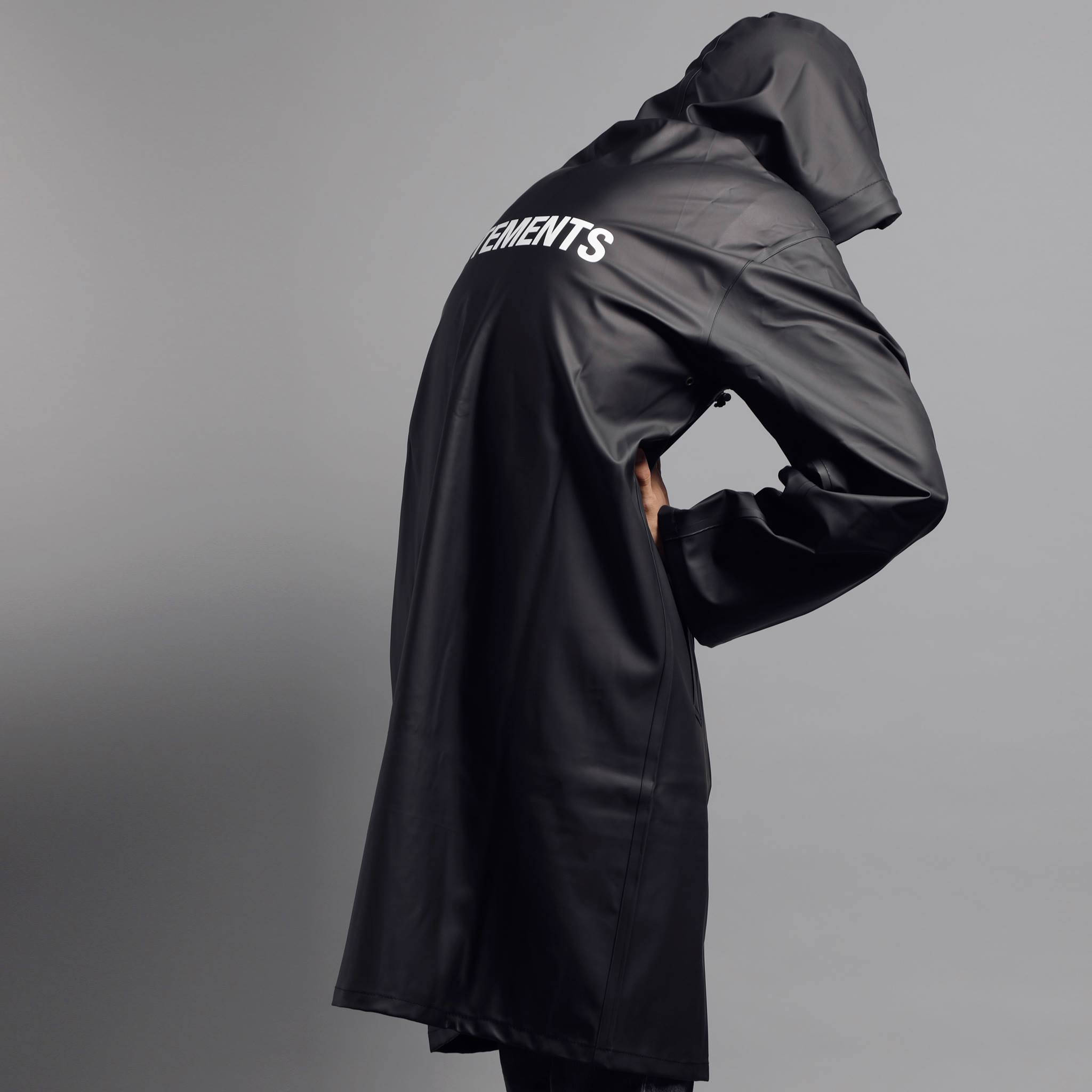 Vetements 桌布 壁紙 — 一個人穿著黑色連帽雨衣，背面有白色文字，站在純灰色背景前。