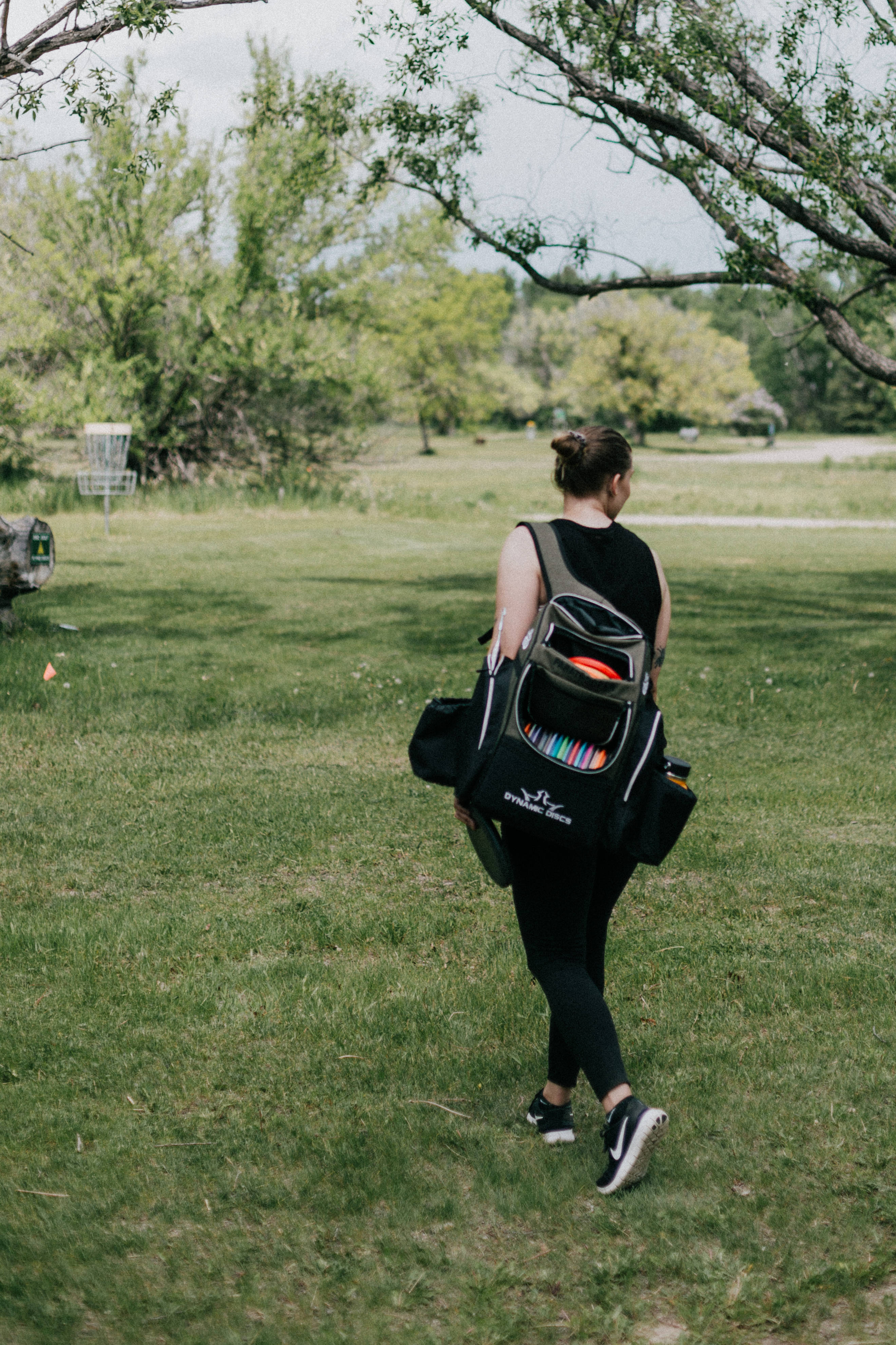Frisbee 桌布 壁紙 — 一個人穿過草地公園，攜帶一個飛盤袋，裡面有色彩繽紛的飛盤可見，正走向一個飛盤高爾夫籃框。