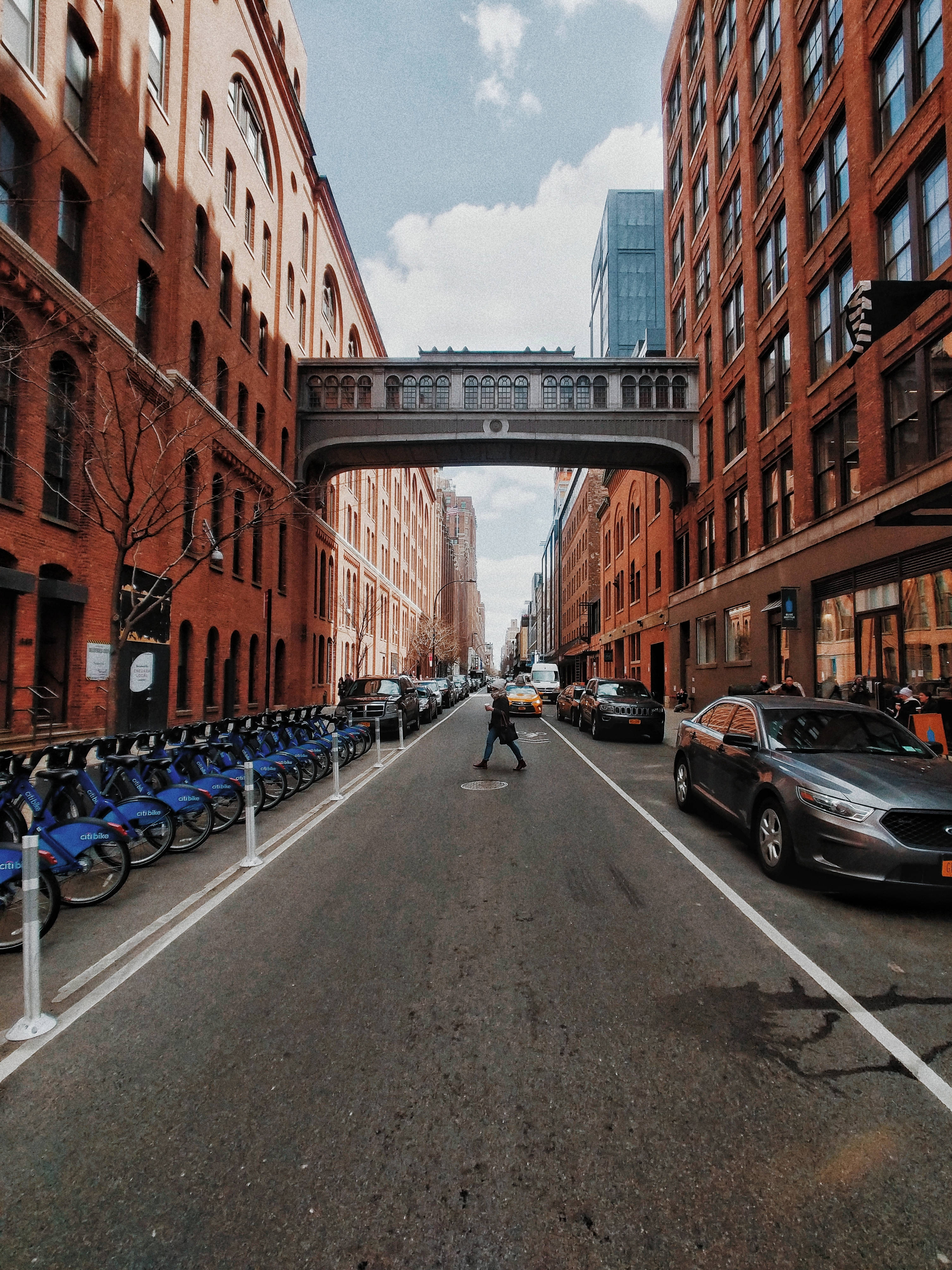 chelsea fc logo 桌布 壁紙 — 一個人穿過陽光普照的城市街道，兩旁是紅磚建築和頭頂上的天橋。