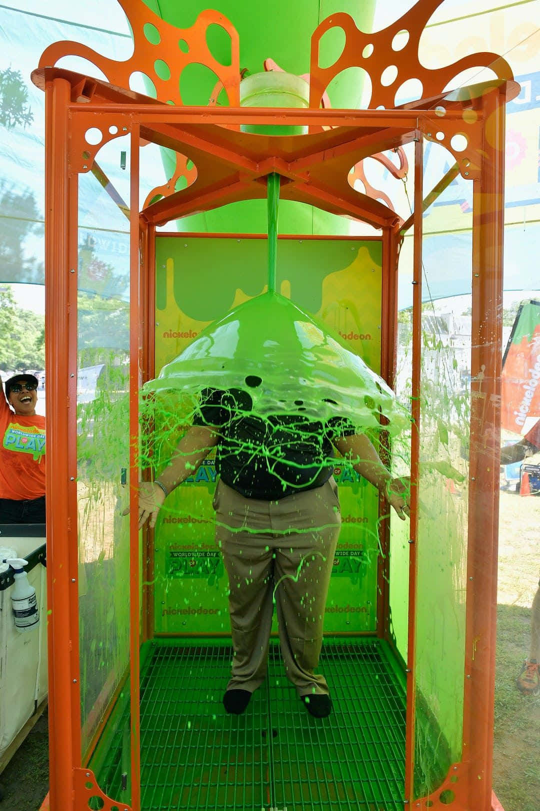 nickelodeon 桌布 壁紙 — 一個人站在一個鮮橙色的黏液大砲攤位內，綠色黏液在戶外活動中濺到他們身上。
