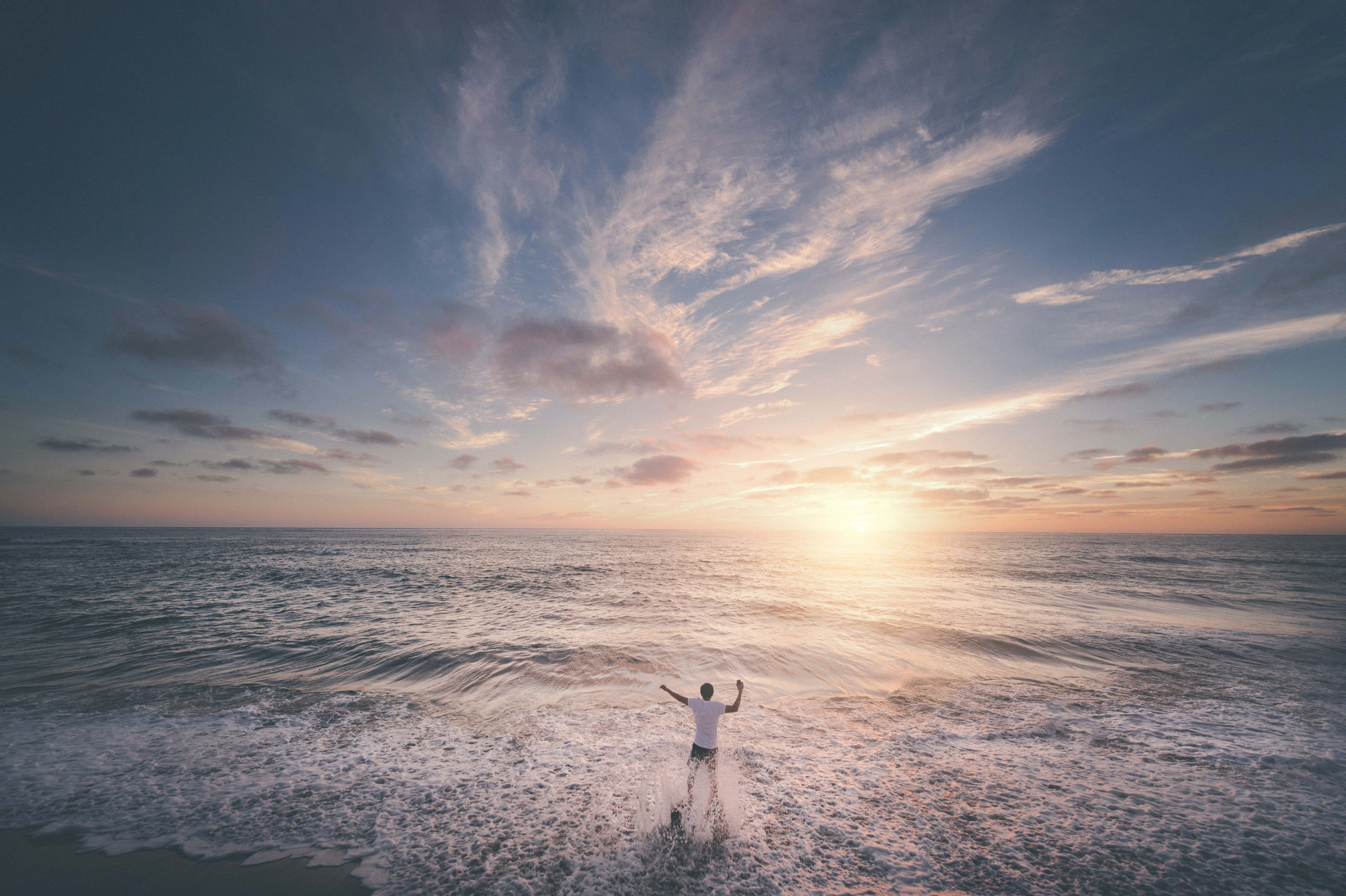 Photography Desktop 桌布 壁紙 — 一個人站在及腰的海浪中，雙臂高舉，夕陽在他們身後落下，將金色光芒灑在水面和天空上。