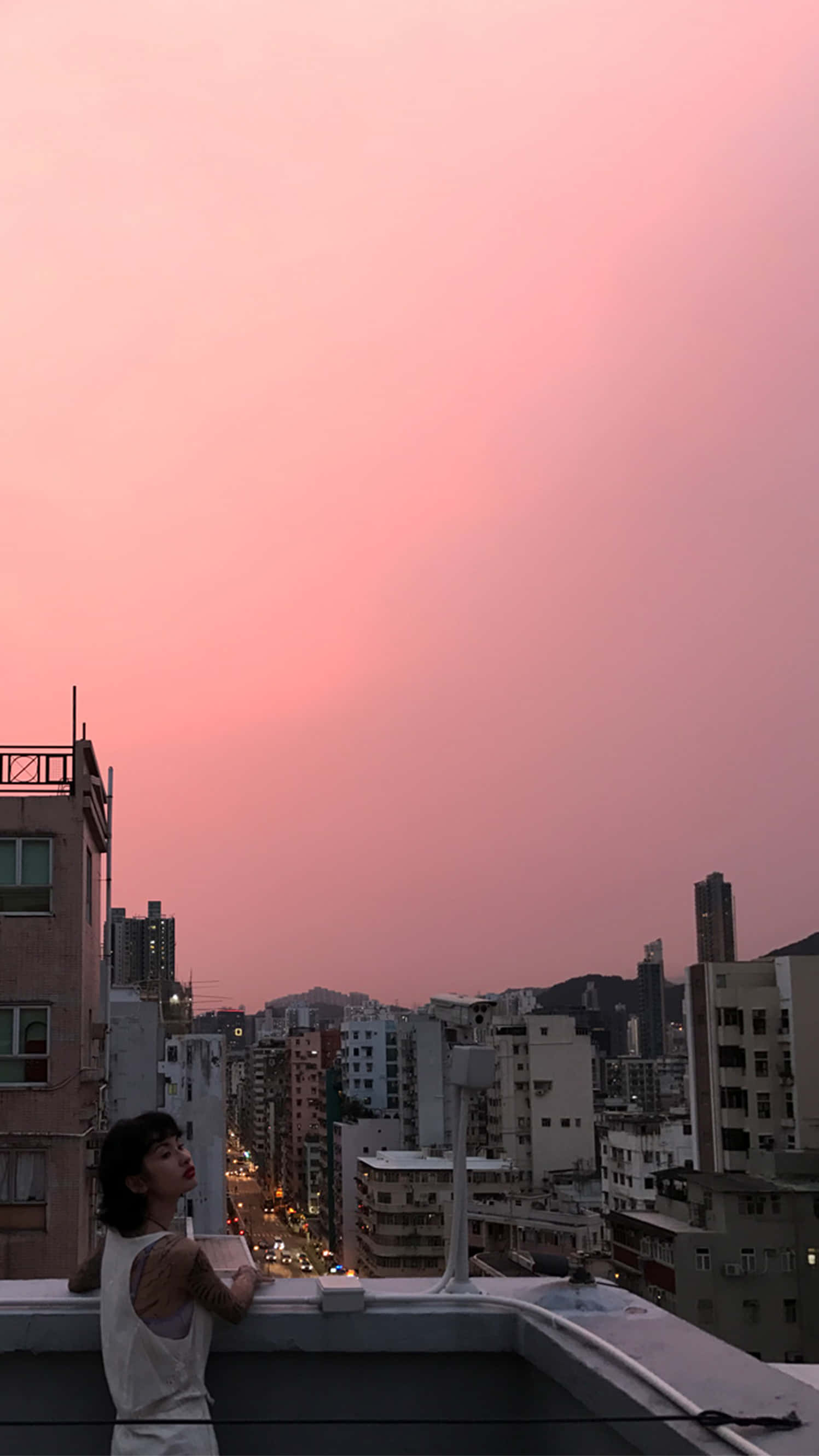 Rooftop 桌布 壁紙 — 一個人站在屋頂上，俯瞰著沐浴在柔和粉紅色日落光中的城市景觀。