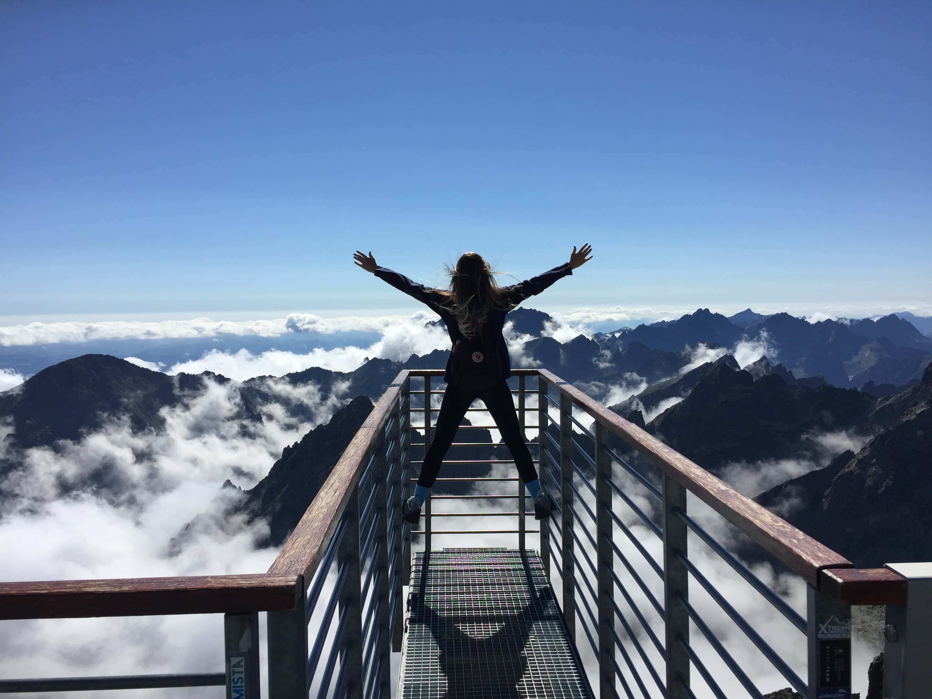 life 桌布 壁紙 — 一個人站在山頂觀景台上，雙臂張開，下方是雲海，遠處是鋸齒狀的山峰，在晴朗的藍天下延伸。
