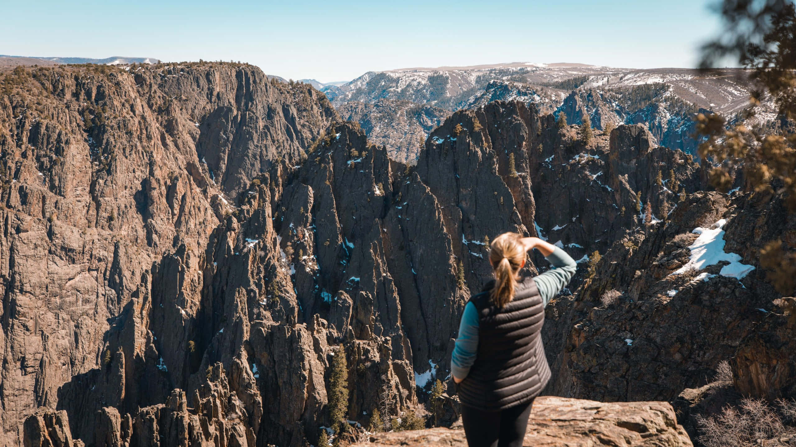 Black Canyon 桌布 壁紙 — 一個人站在岩石懸崖邊，俯瞰廣闊崎嶇的峽谷，白雪覆蓋的山峰在晴朗藍天下。
