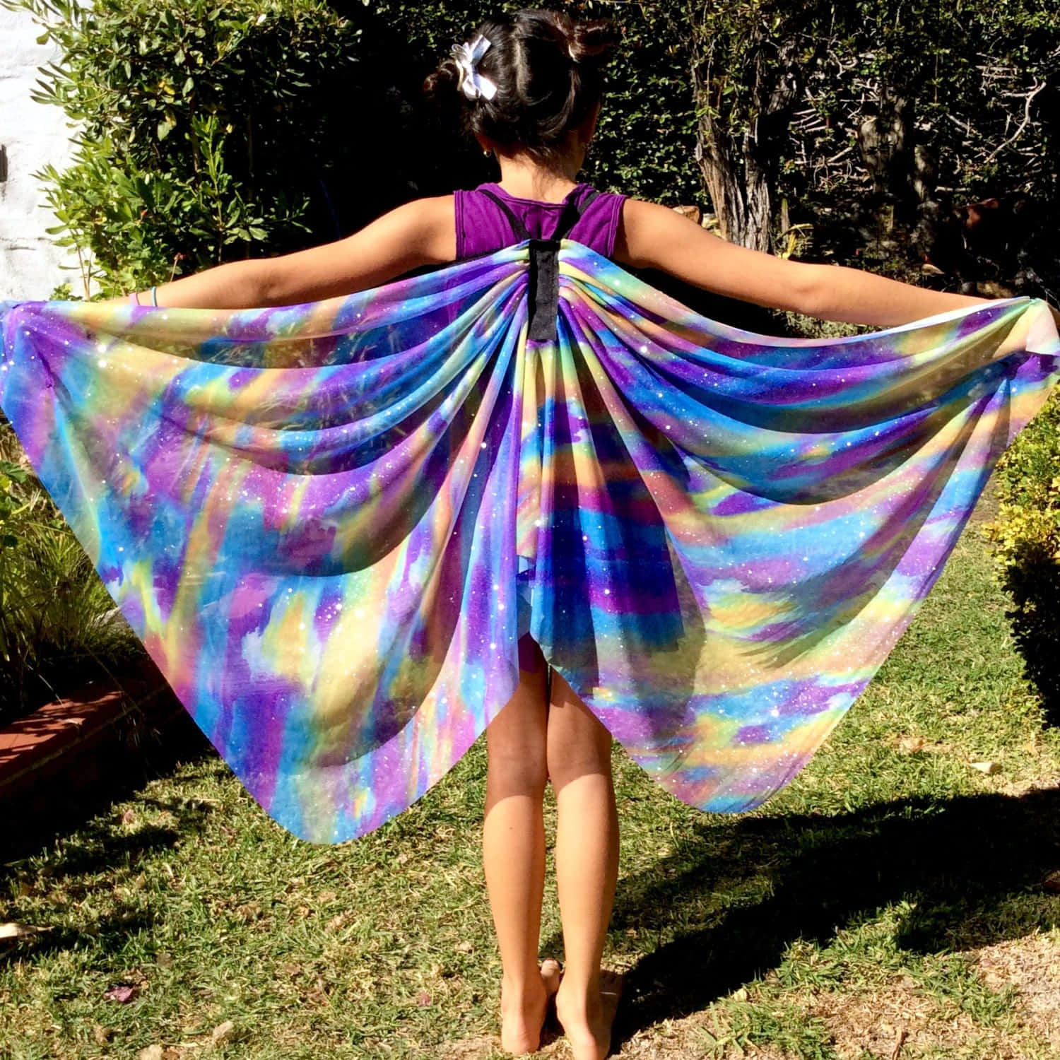 Butterfly Wing Dress 桌布 壁紙 — 一個人站在戶外，雙臂張開，穿著鮮豔的銀河圖案斗篷，在陽光普照的花園背景下像宇宙翅膀般飄揚。