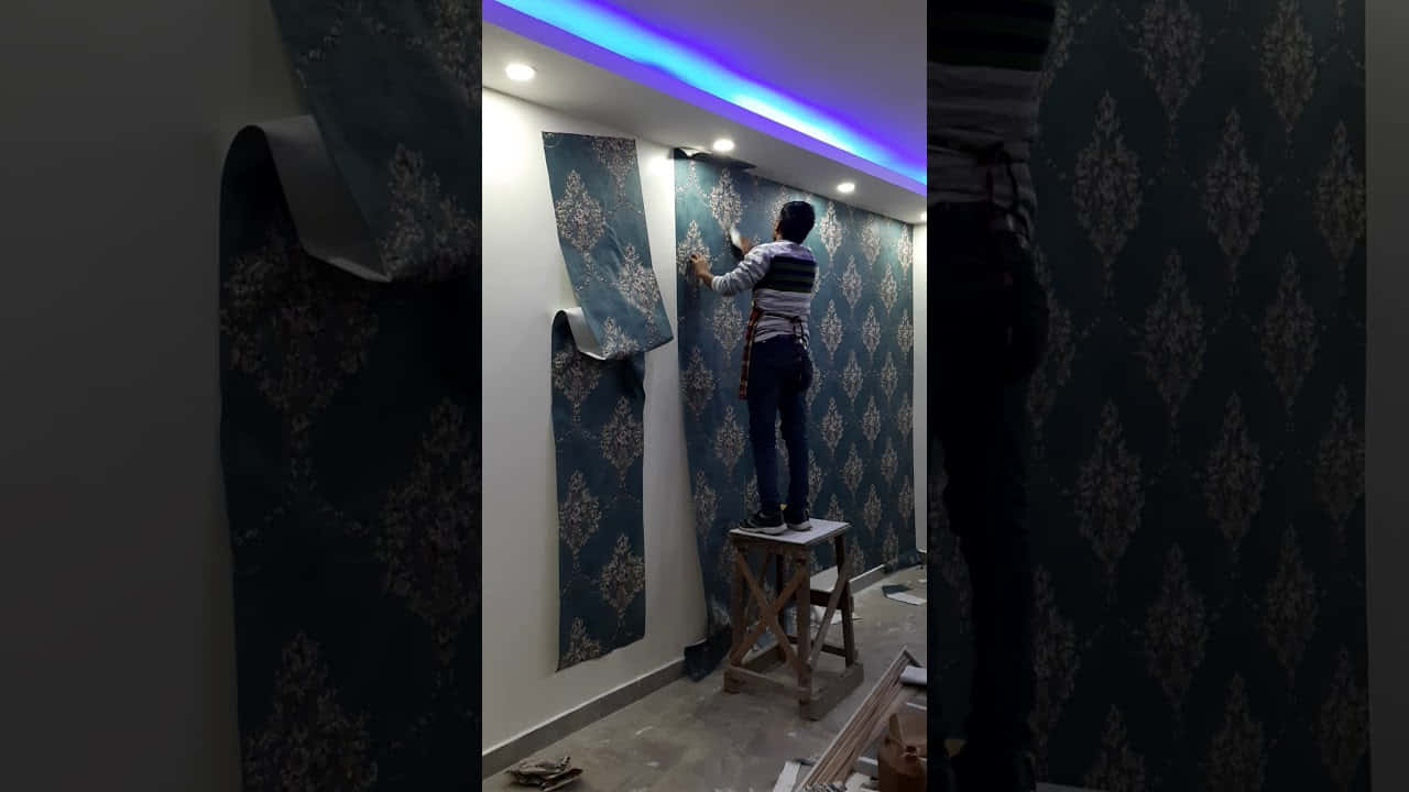 Installation Art 桌布 壁紙 — 一個人站在木凳上，在閃爍的藍色LED燈光下將深藍色大馬士革花紋壁紙貼在牆上。